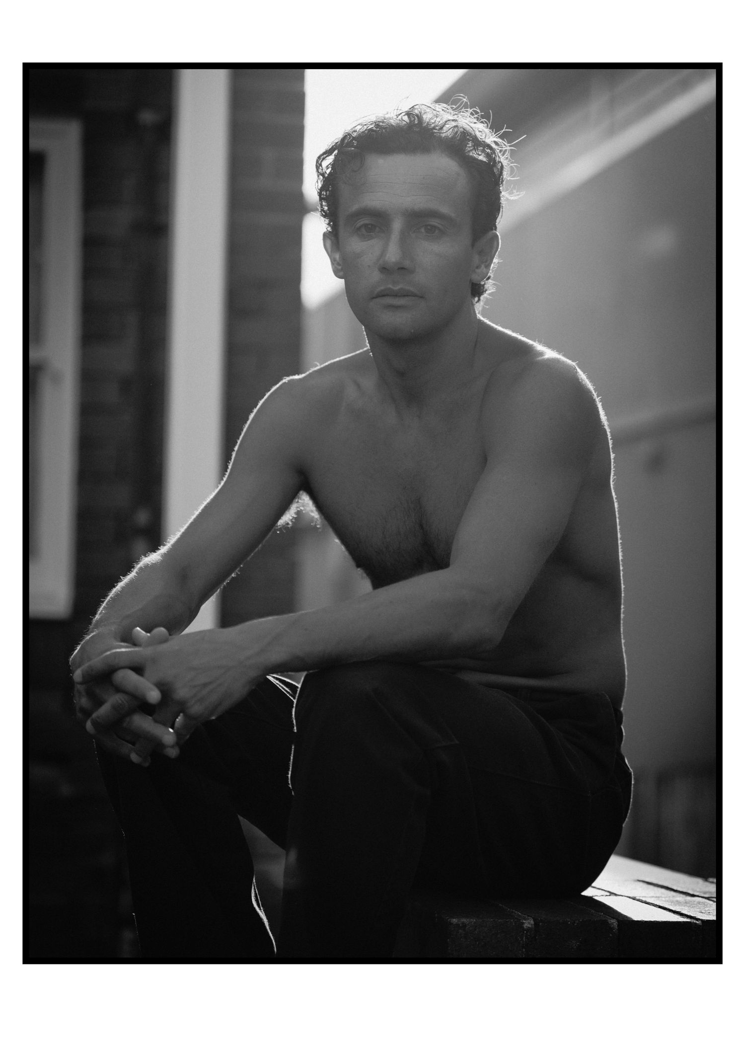 Shirtless man with curly hair sitting pensively in dramatic black and white portrait.