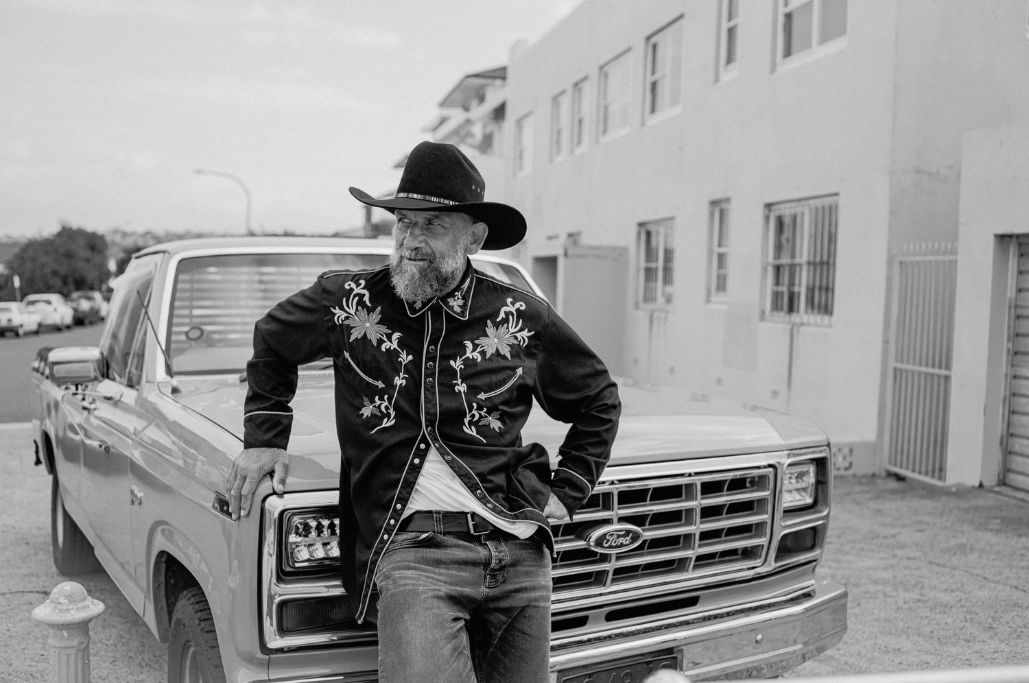 Man in cowboy hat and embroidered western shirt leaning against vintage Ford pickup truck, black and white photo.