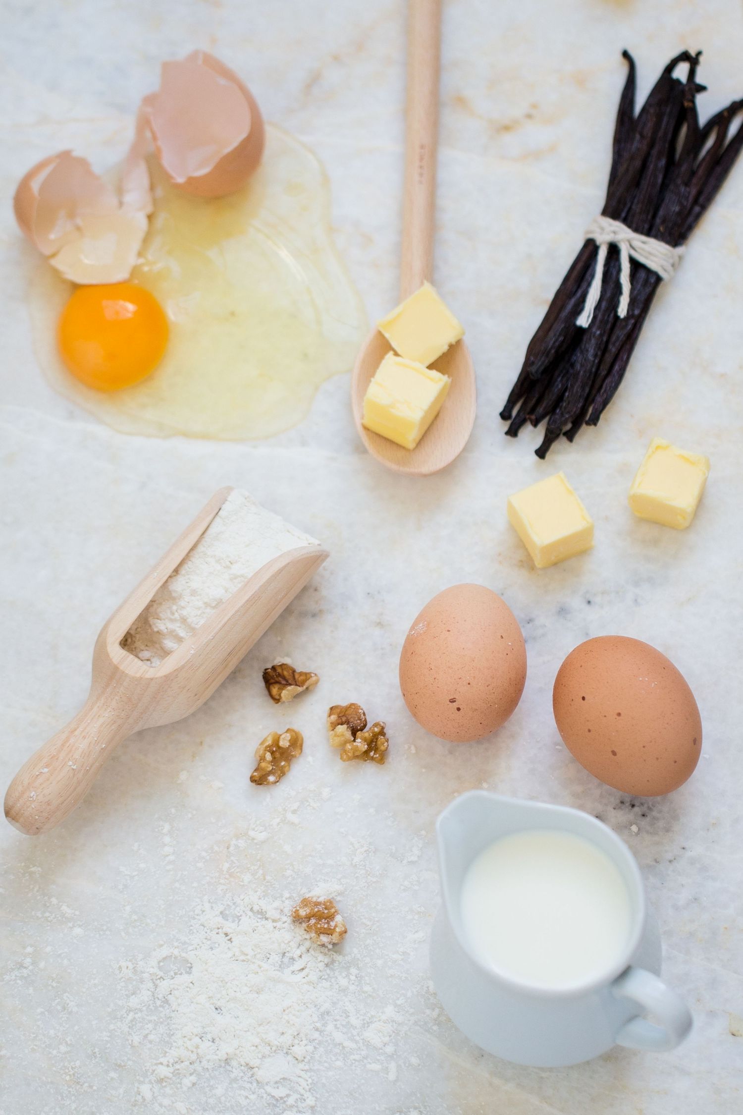 Baking ingredients flatlay with eggs, butter cubes, vanilla beans, walnuts, and milk on marble surface.
