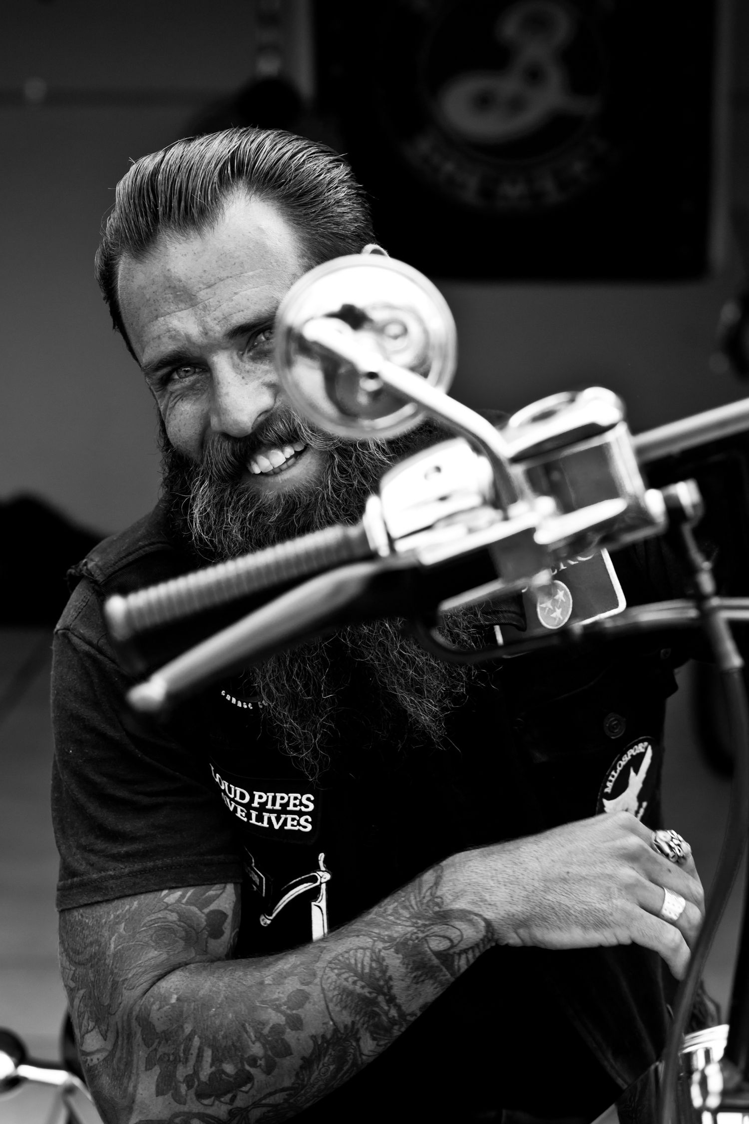 Bearded tattooed man smiling behind motorcycle handlebars in black and white.