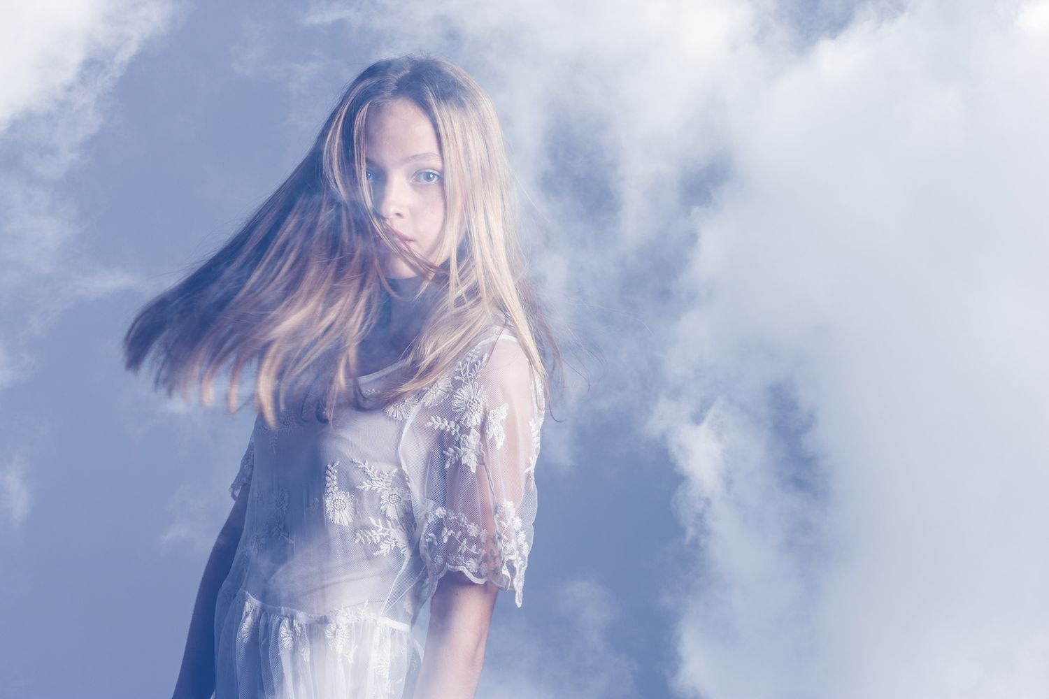 Ethereal blonde woman in white lace dress surrounded by soft clouds in dreamy double exposure.