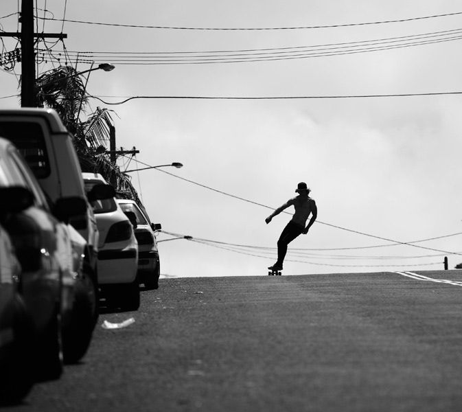 Silhouette of a skateboarder riding downhill on a street lined with parked cars.