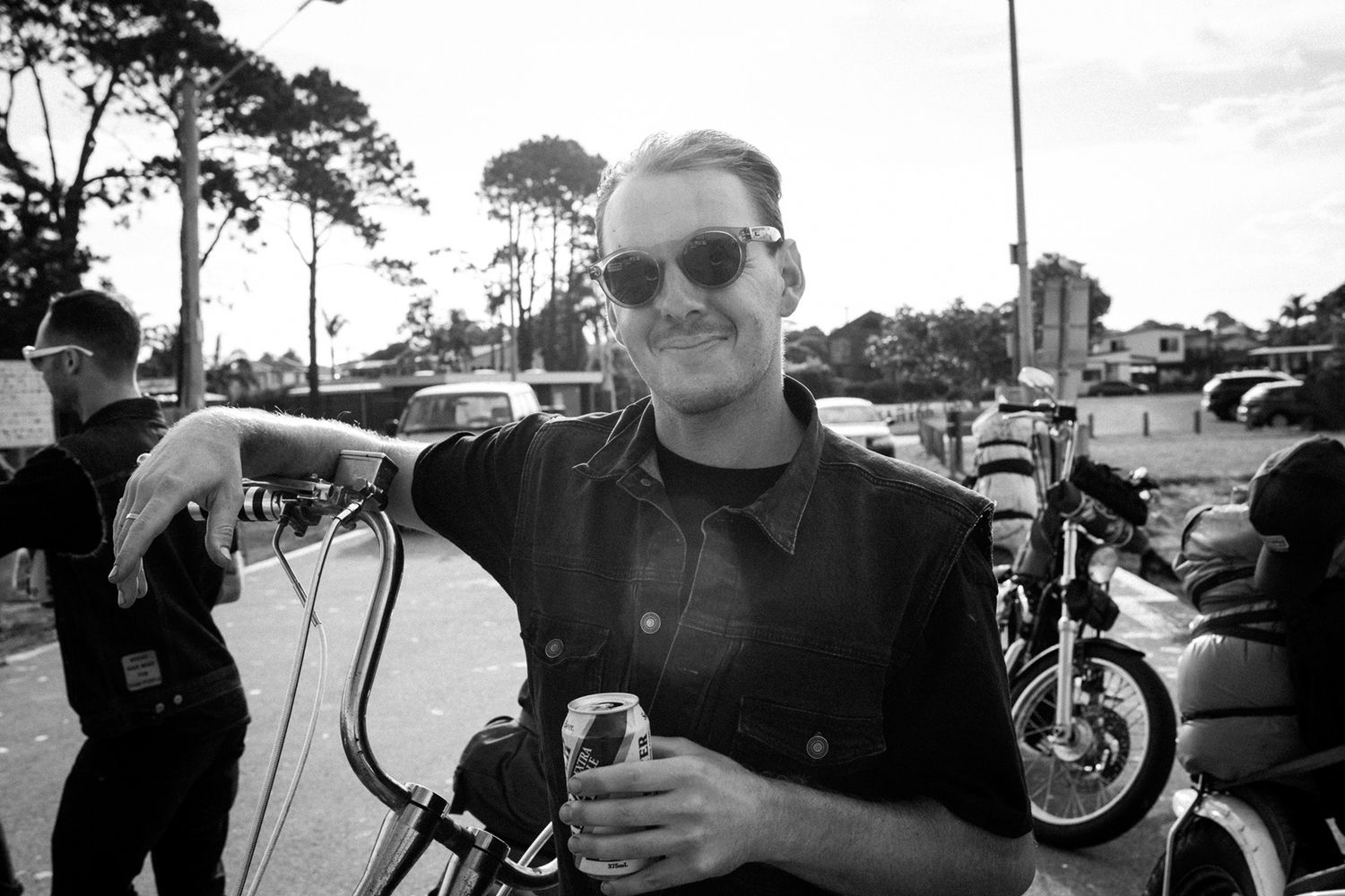 Black and white photo of a smiling man in sunglasses leaning on a motorcycle at an outdoor event.