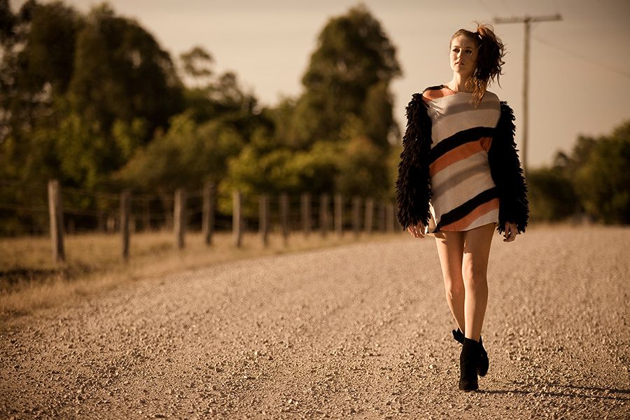 Young woman in striped dress and black fur coat walking down a rural gravel road at sunset.