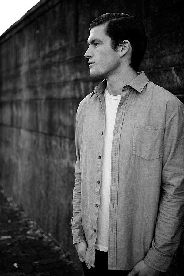 A black and white portrait of a young man in a button-up shirt standing against a stone wall.