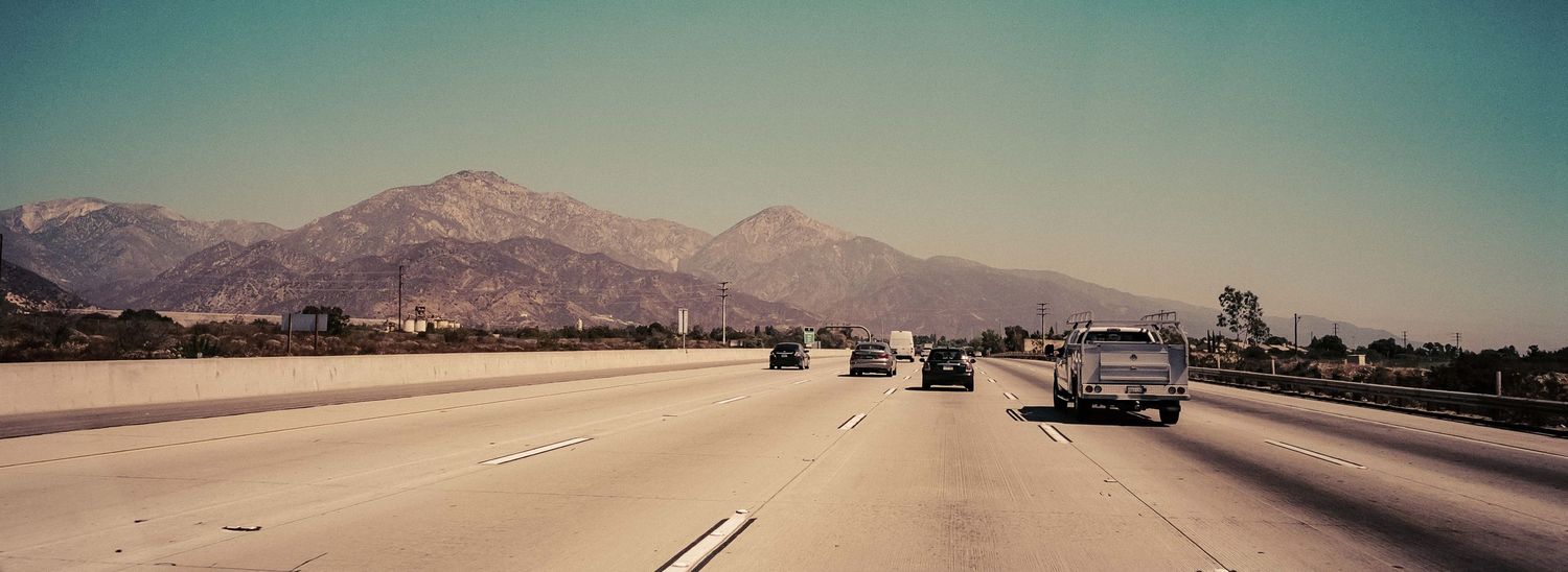 Vintage panoramic view of cars driving on wide California freeway with snow-capped mountains in background.