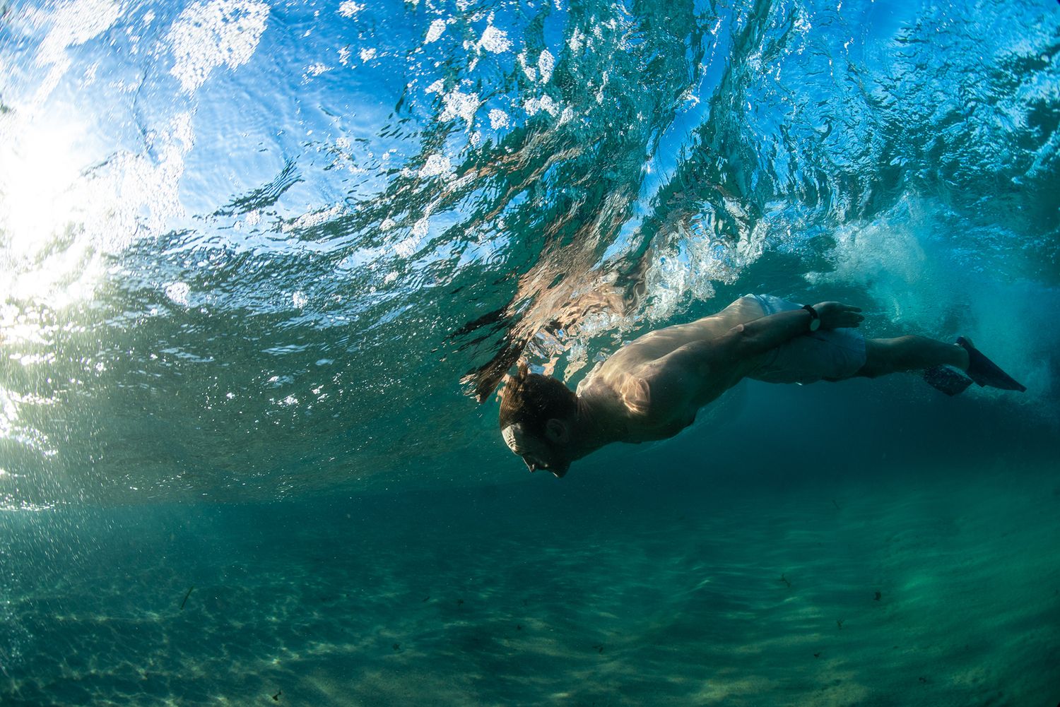 Underwater view of a person swimming beneath a breaking ocean wave in clear blue water.