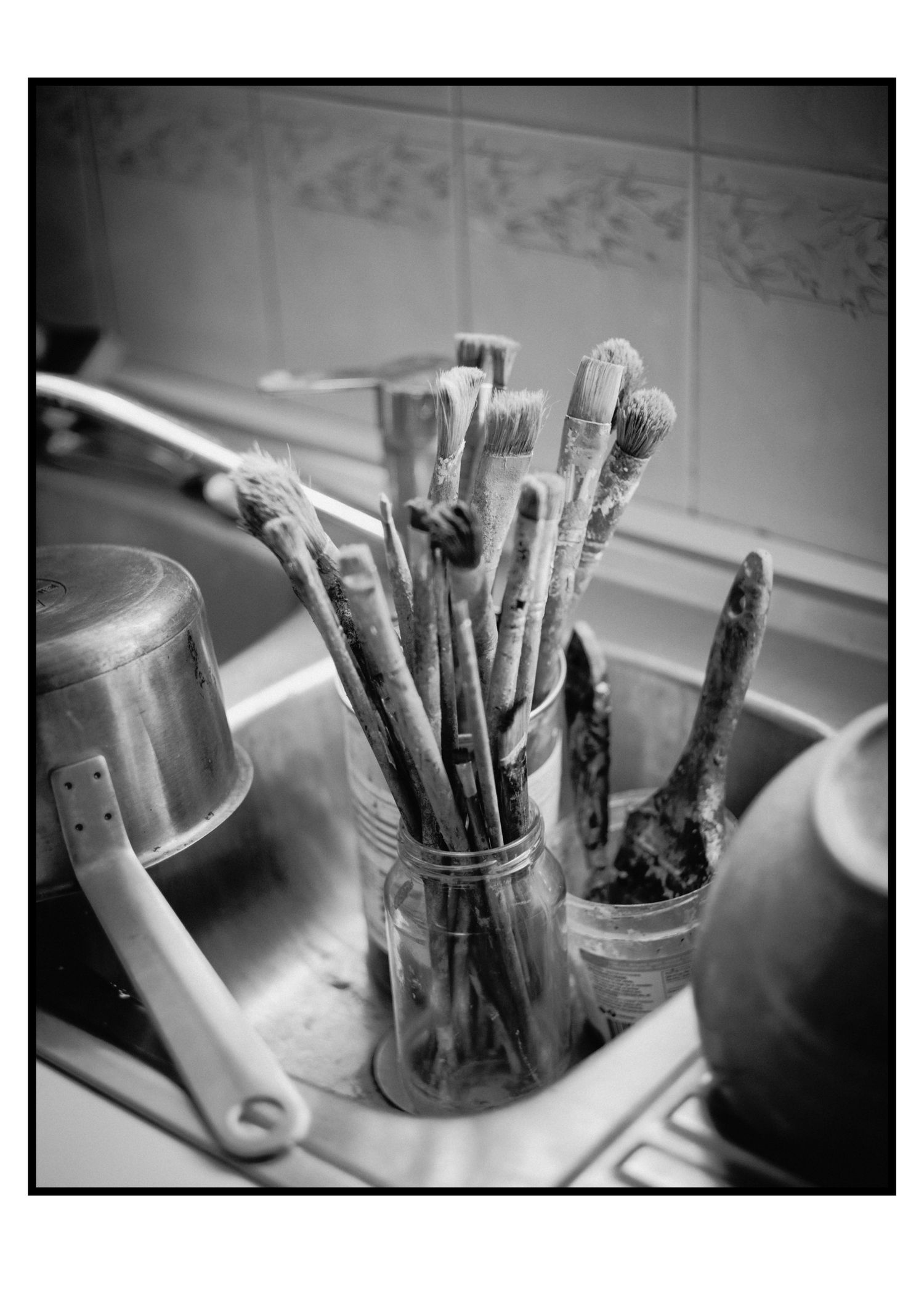 Black and white photo of paint-covered brushes stored in glass jar beside kitchen sink.