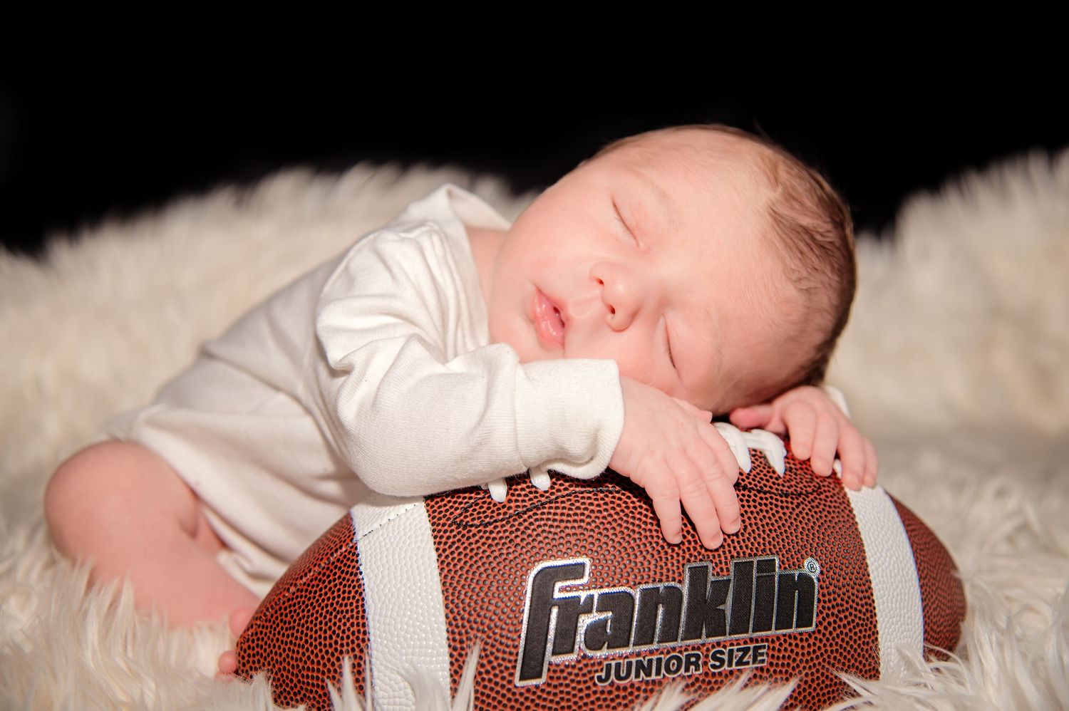 Newborn baby sleeping peacefully on soft cream-colored blanket with football prop.