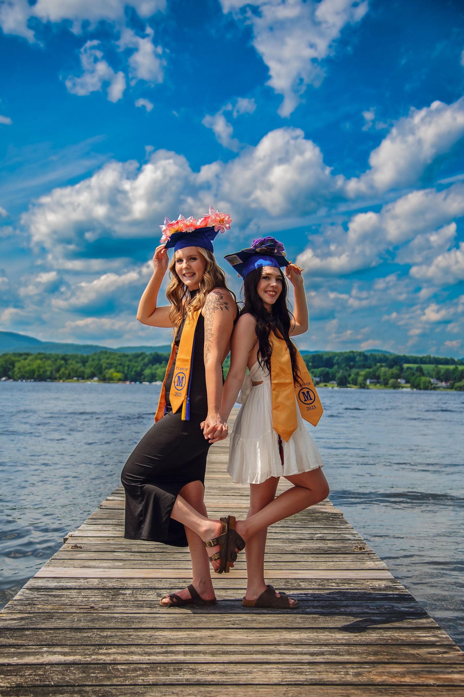 Graduates celebrate on a lakeside dock with dramatic blue sky and puffy clouds in the background.