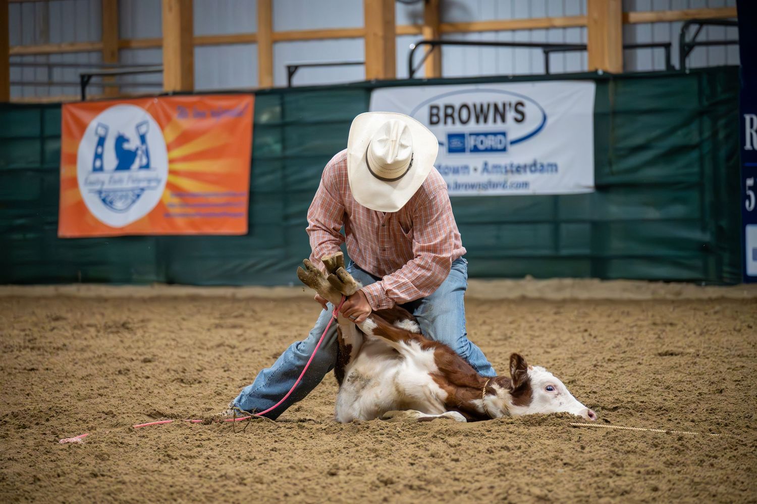 Rodeo rider attempts to rope a calf in indoor arena with Brown's banner visible.