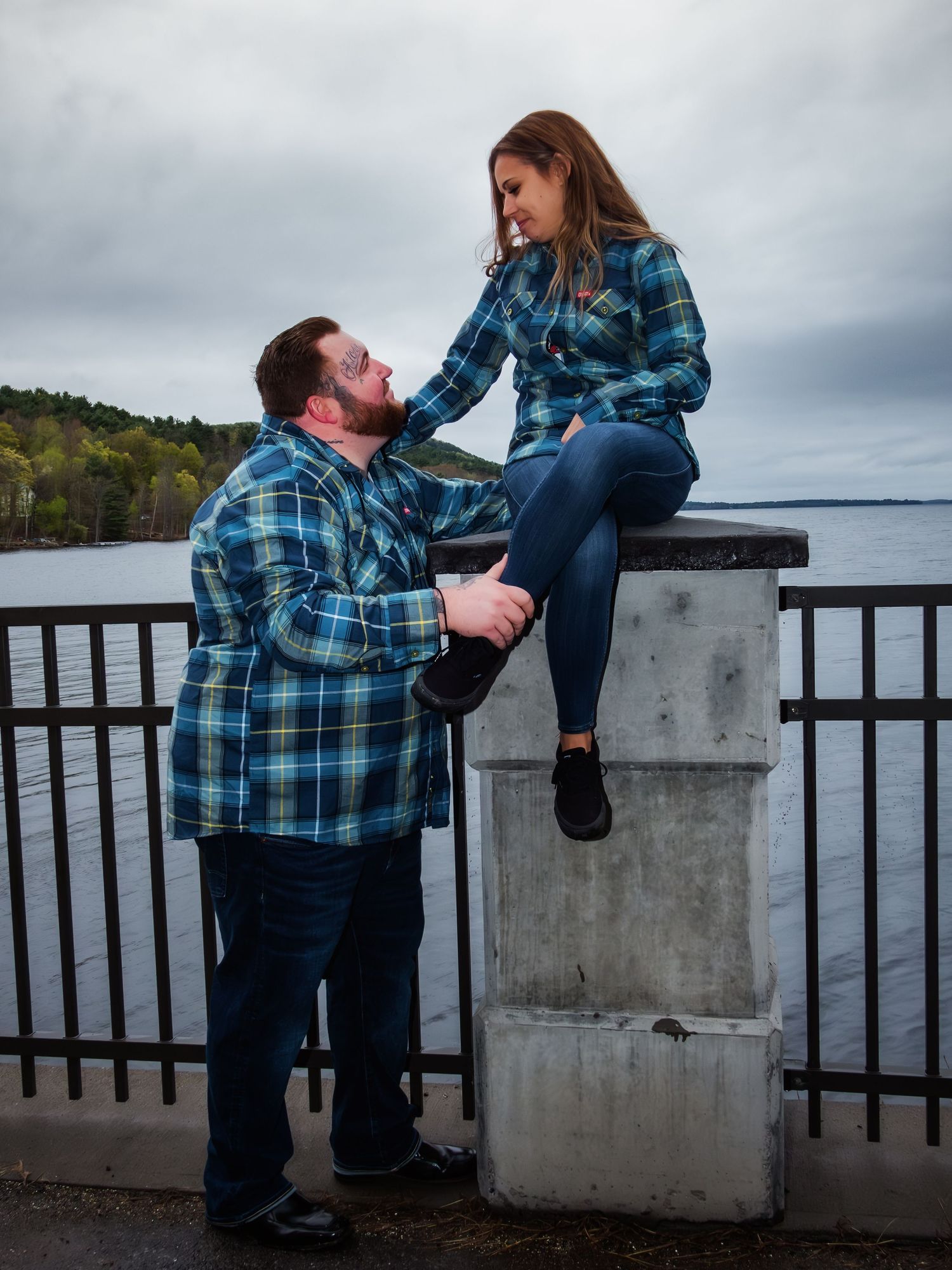 A couple shares a romantic moment on a waterfront boardwalk overlooking a scenic lake on an overcast day.