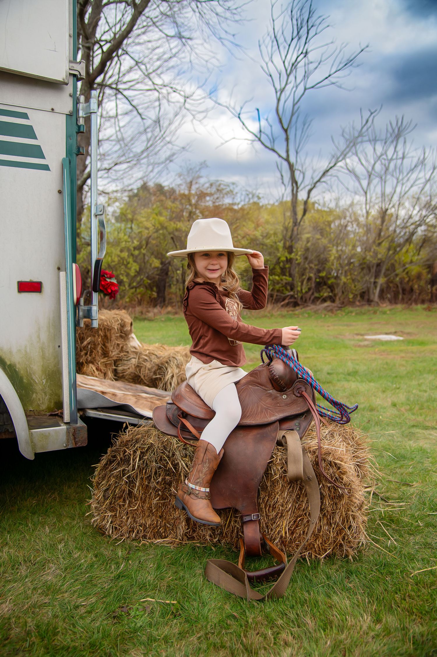 A western-style saddle rests on a hay bale in a grassy field next to a white trailer.