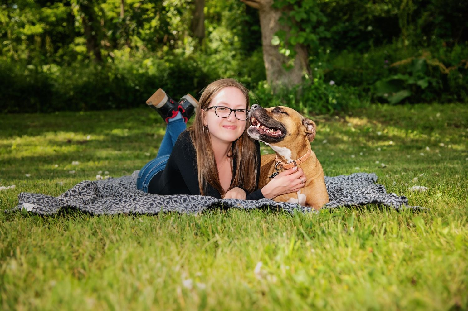 A dog and companion relax together on a blanket in sunny green grass.