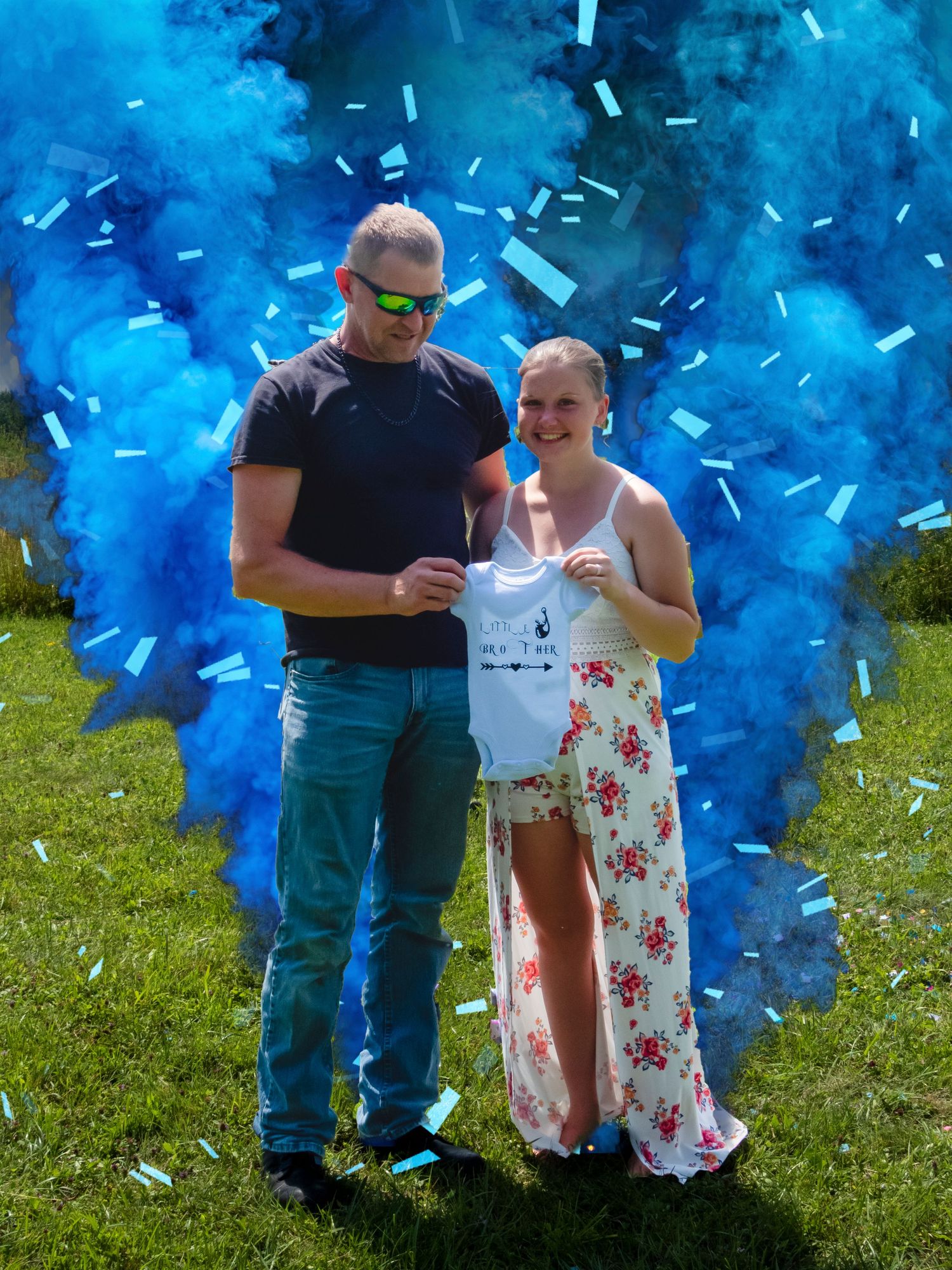 A couple stands amid blue smoke and confetti during an outdoor gender reveal celebration.