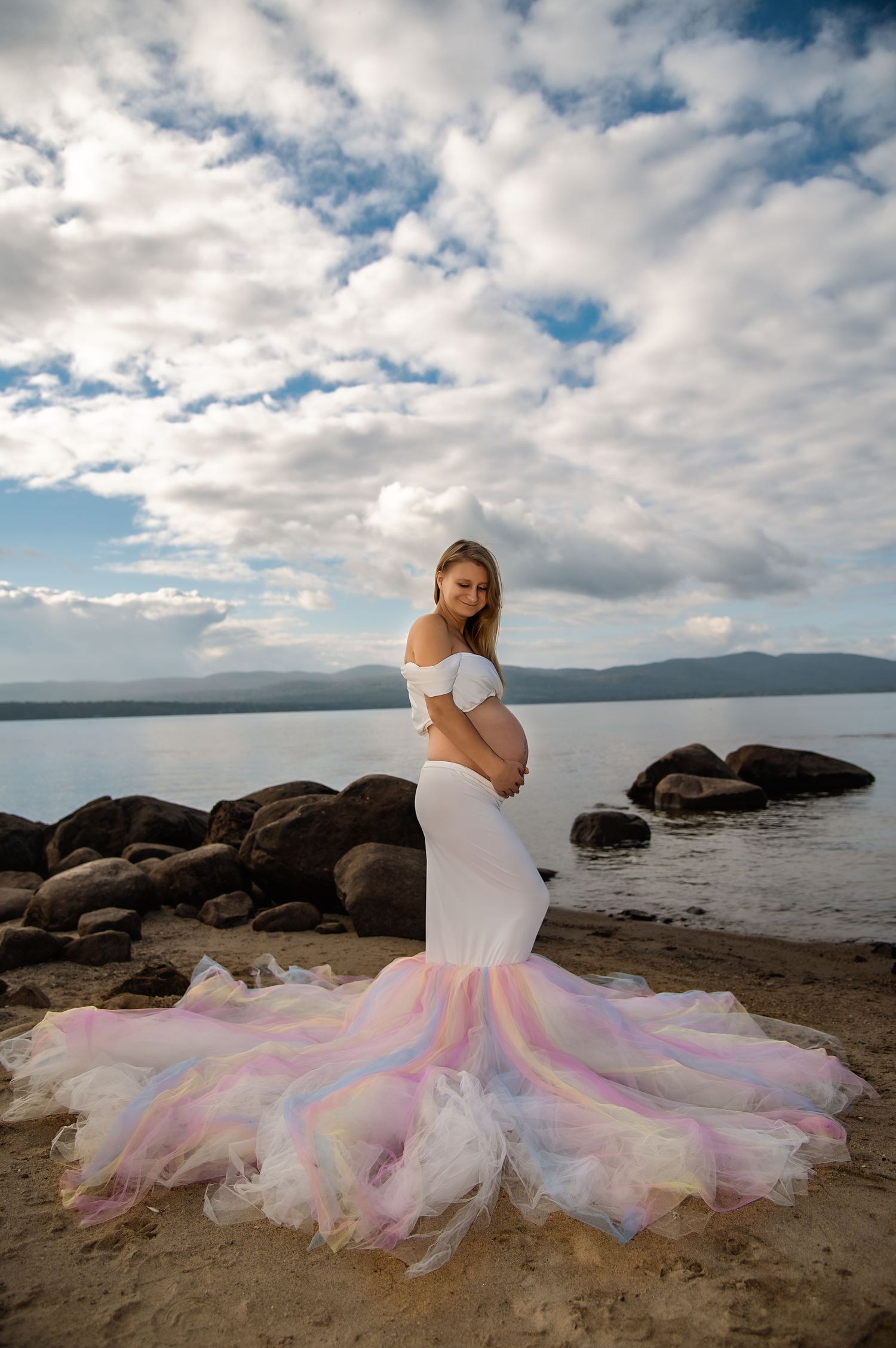 Woman in white dress with colorful tulle skirt poses on rocky beach landscape.