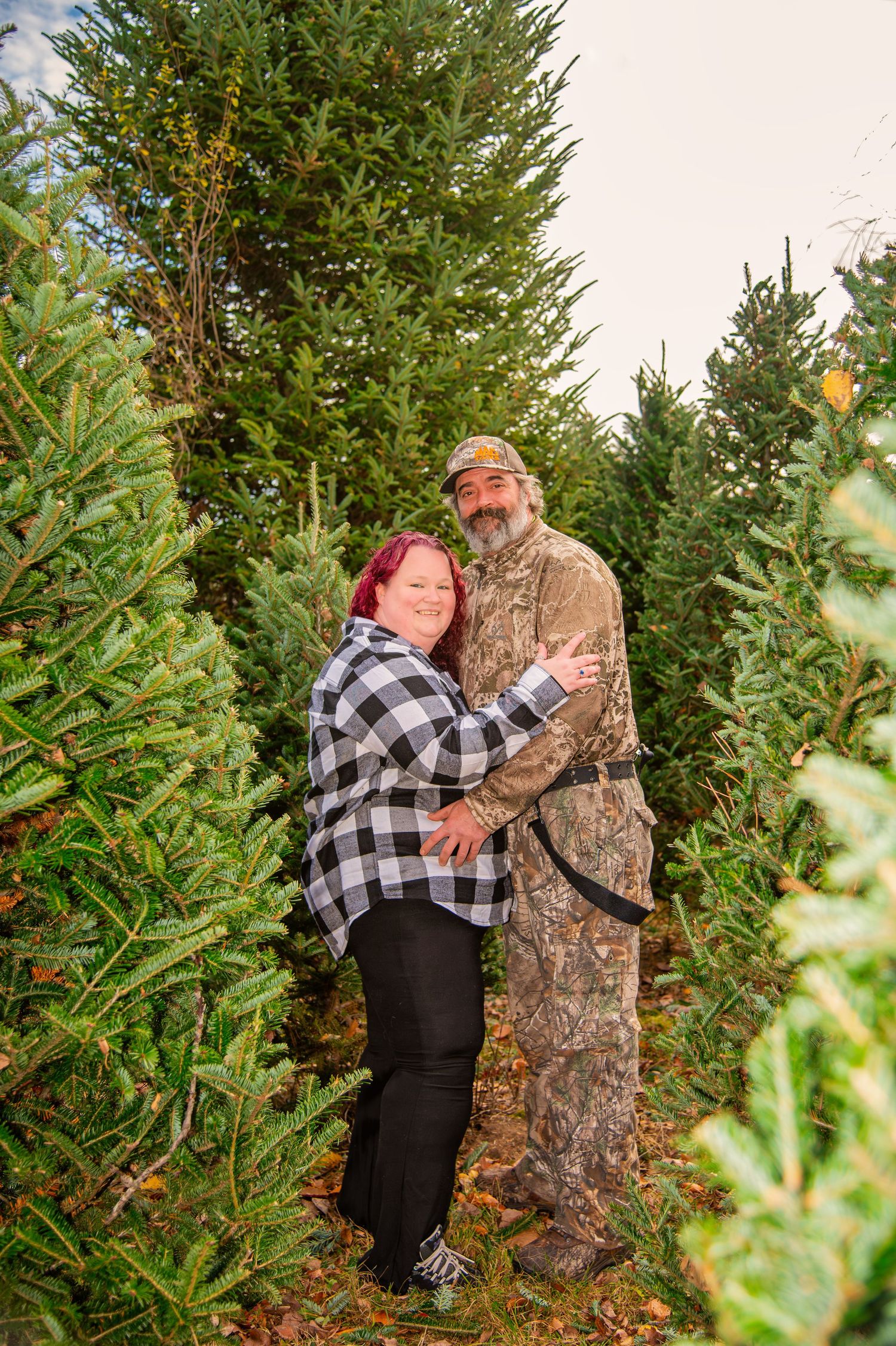 Couple embracing between evergreen trees at a Christmas tree farm.