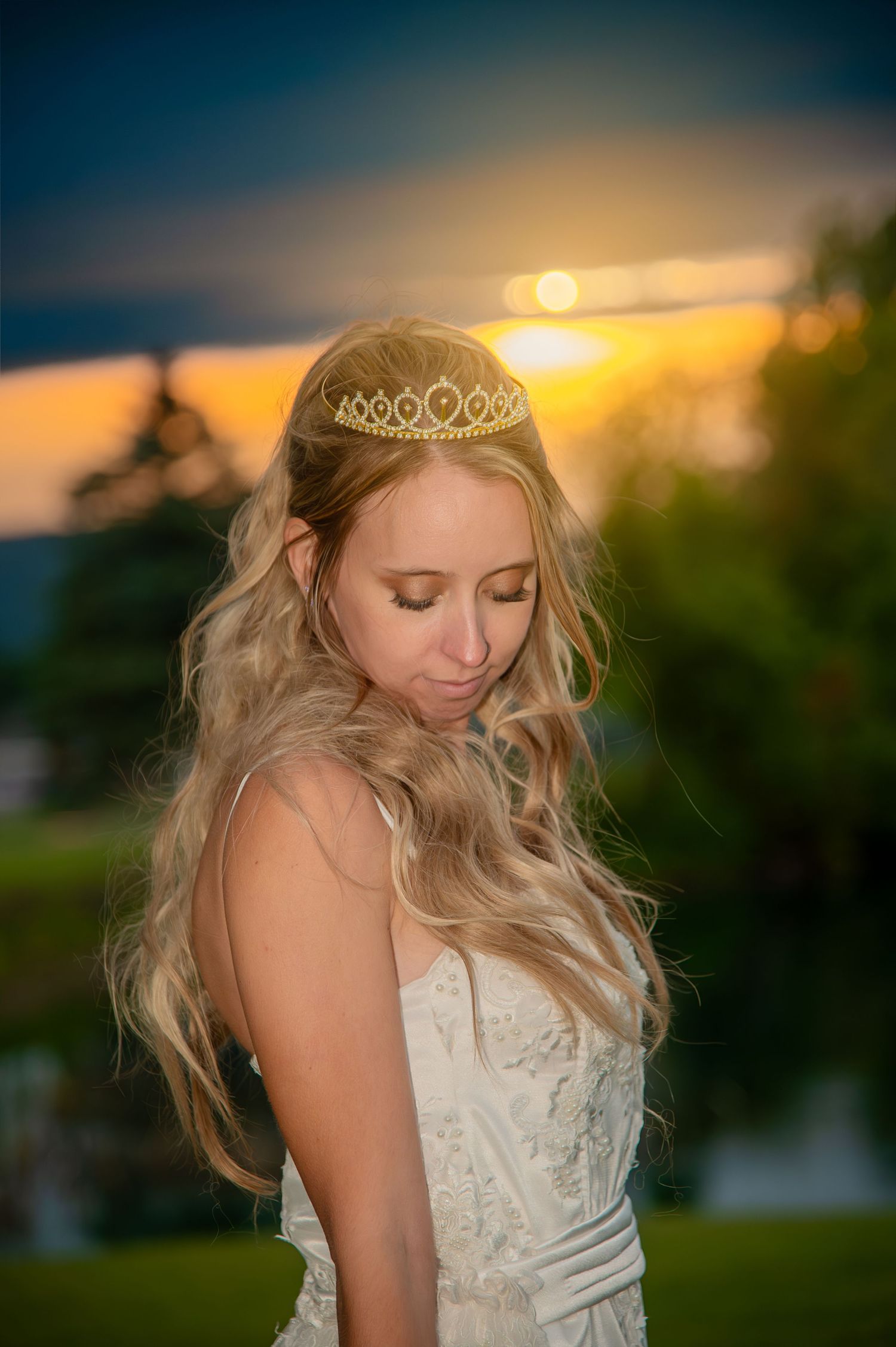 Bride in white dress and tiara poses at golden sunset with dreamy lighting.