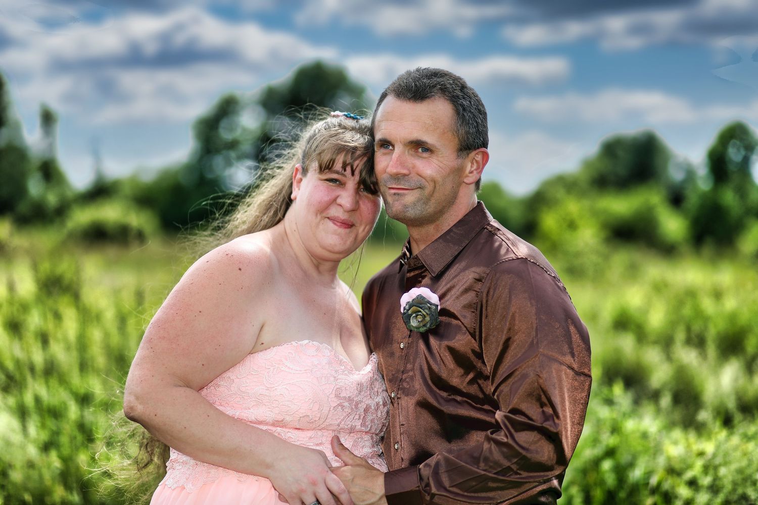 Romantic outdoor portrait of a couple in pink dress and brown shirt against a green field background.