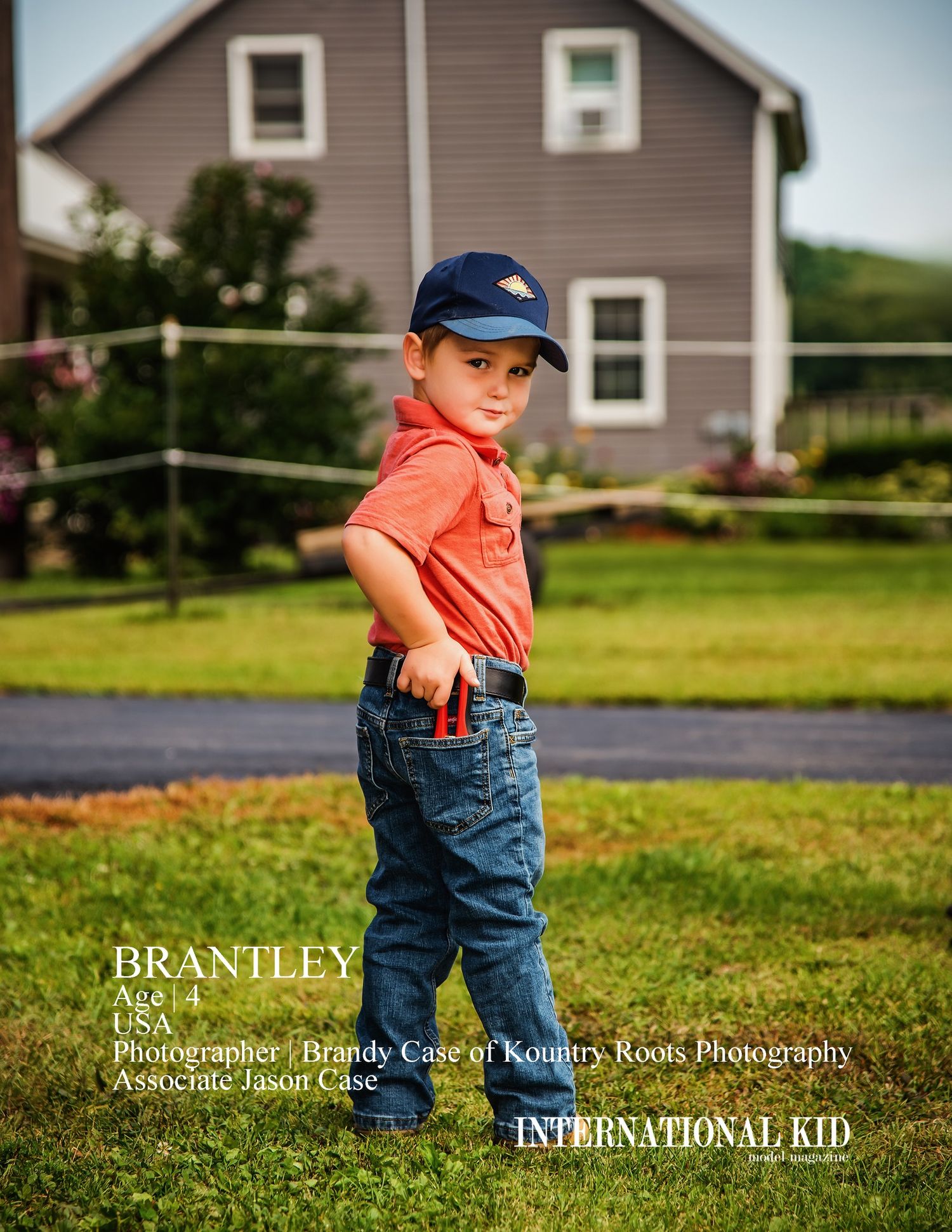 A small child in orange shirt and jeans stands in front of a gray house on a summer day.