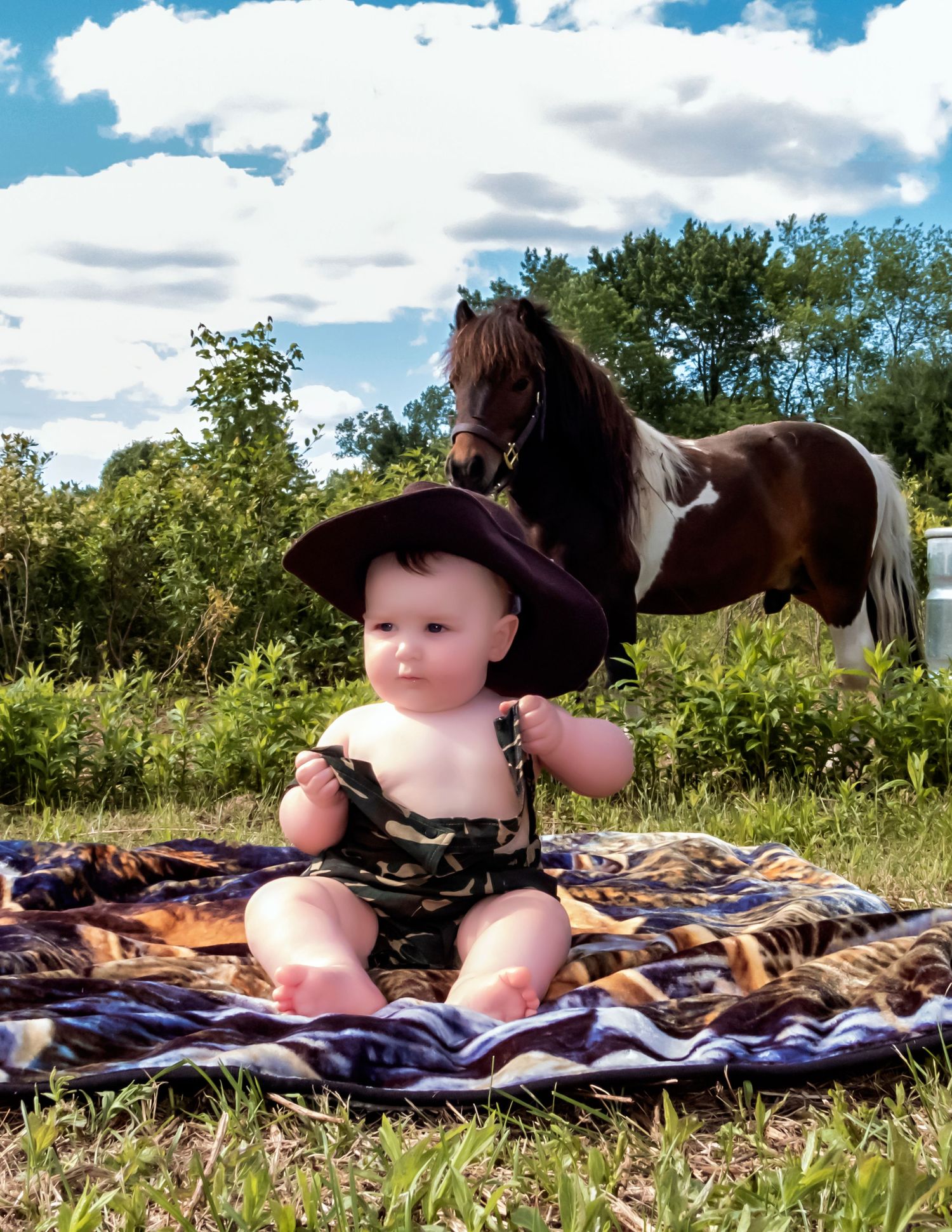 A baby in a cowboy hat sits on a blanket with a paint horse grazing in the background.