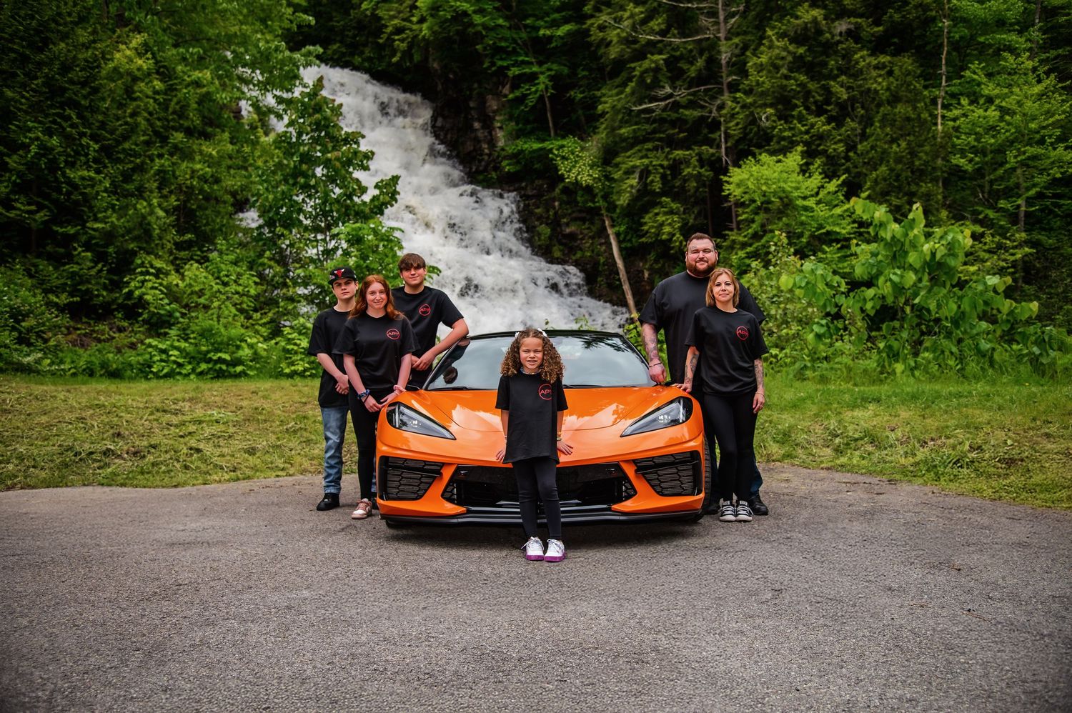 Group photo with orange sports car against scenic waterfall backdrop.