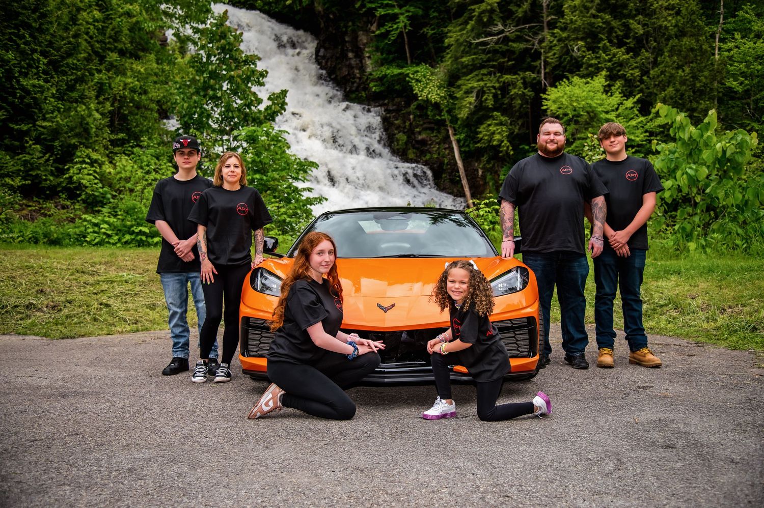 A group poses with orange and black sports cars in an outdoor setting surrounded by green foliage.