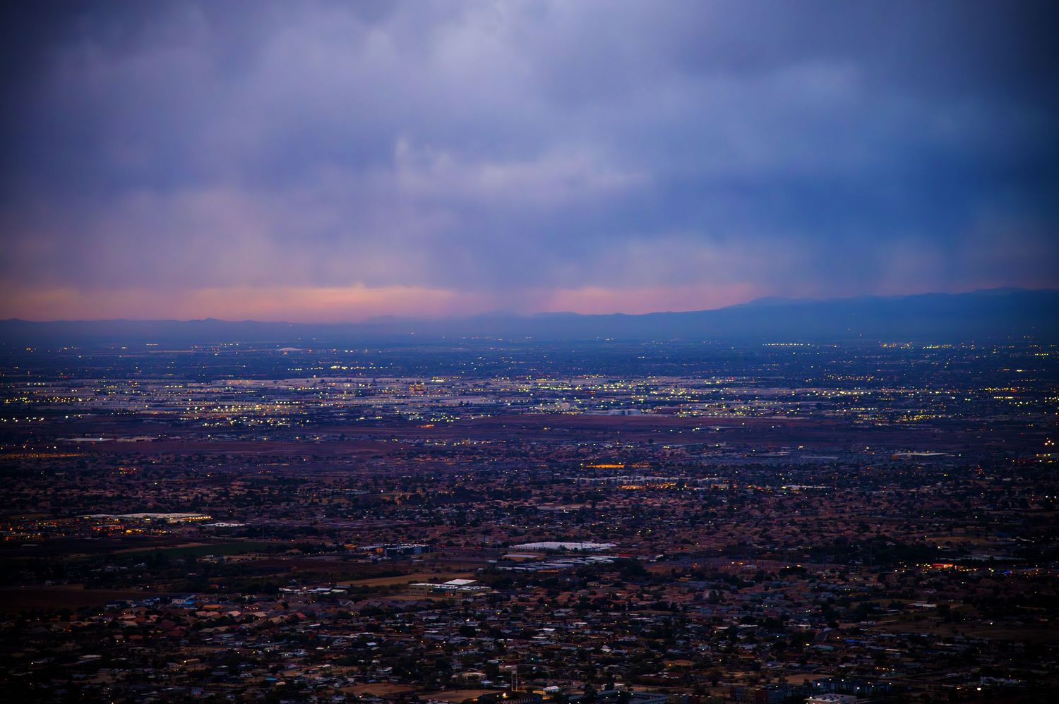 Panoramic cityscape view at dusk showing urban sprawl extending to distant mountains.