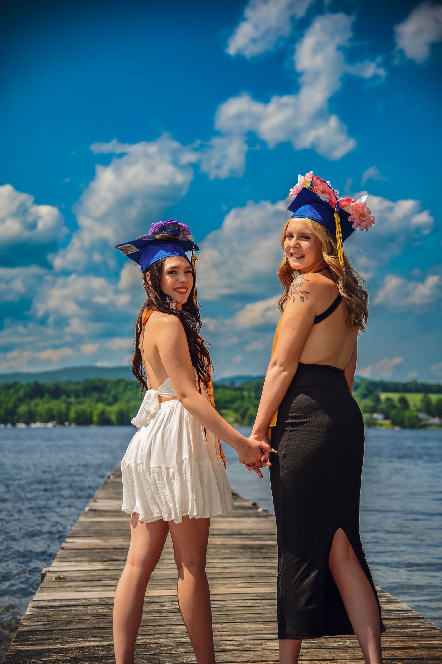 Two graduates in caps and gowns pose playfully on a wooden dock by a lake on a sunny day.
