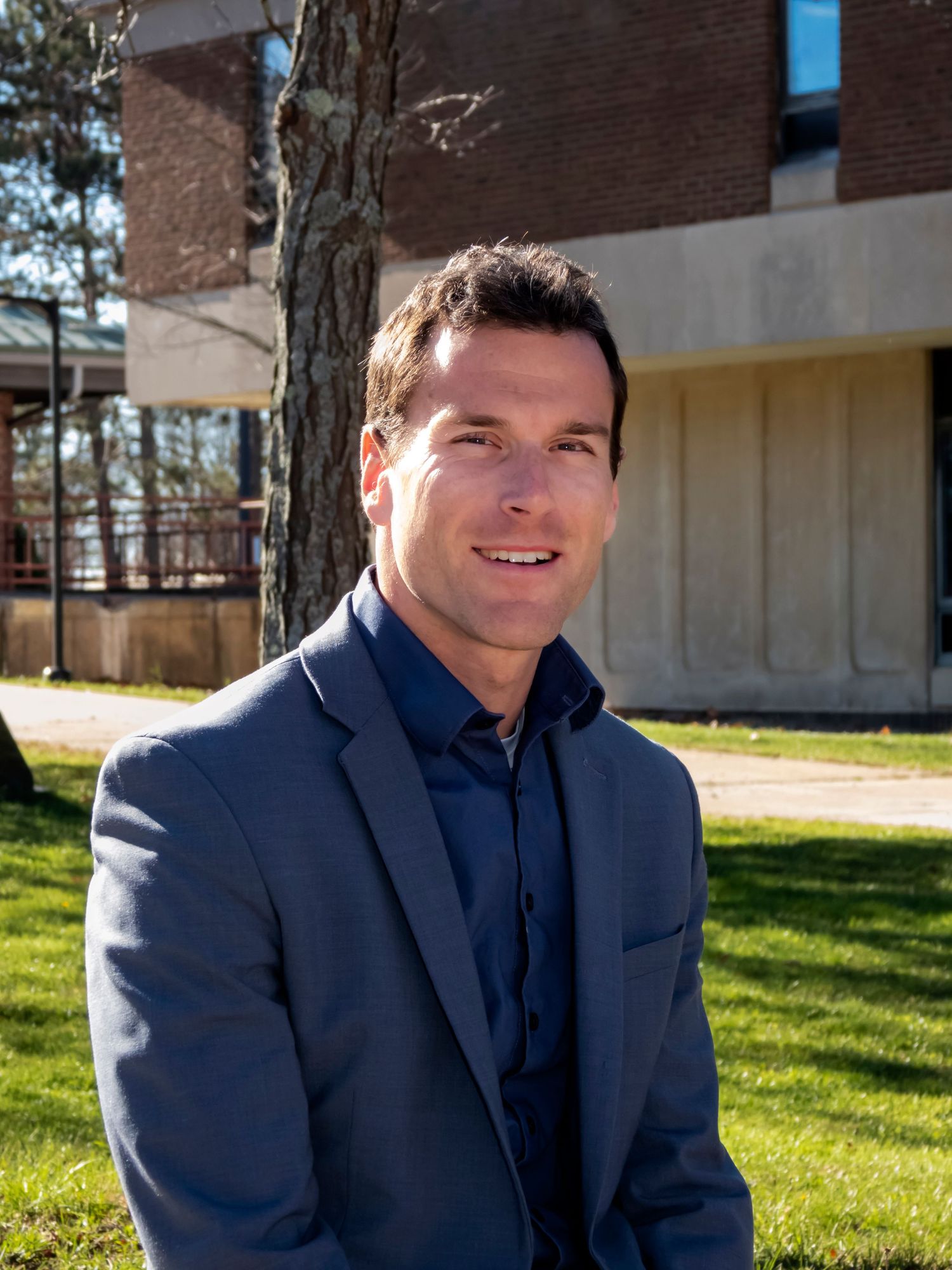 Professional headshot of person in navy suit sitting outdoors on sunny day.