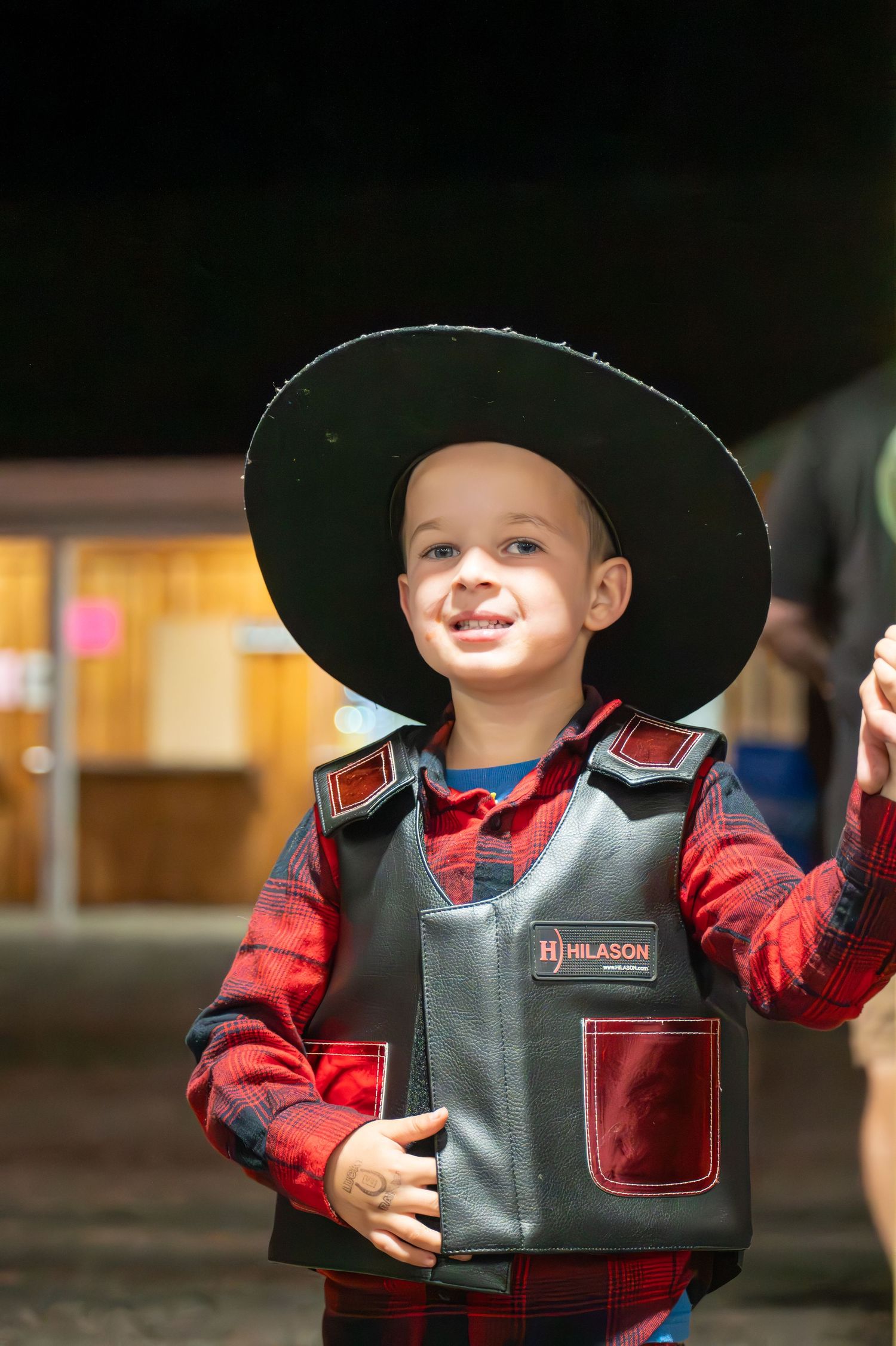 Young child wearing a black cowboy hat and red plaid shirt with leather vest smiles brightly.