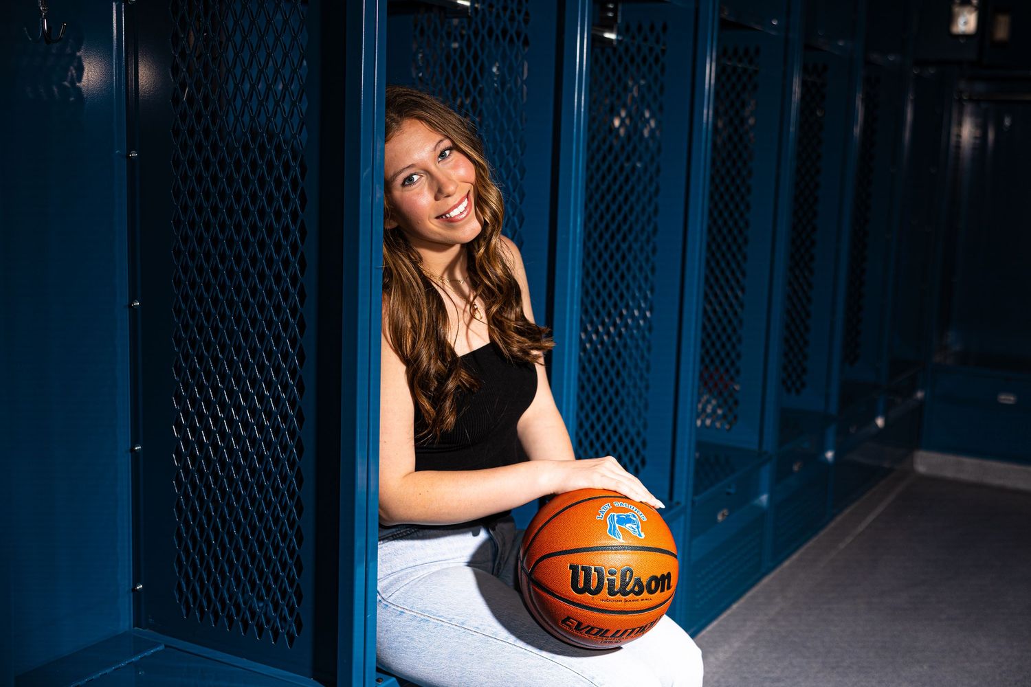 Teenage girl holding a Wilson basketball while sitting in front of blue lockers.
