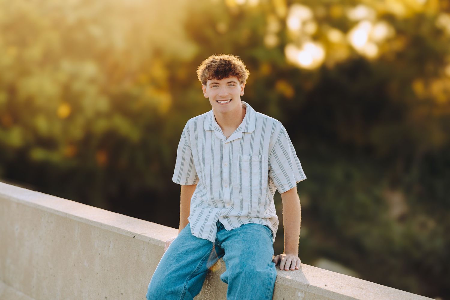 Smiling teenage boy with curly hair sits on a concrete ledge outdoors in golden sunlight wearing jeans.