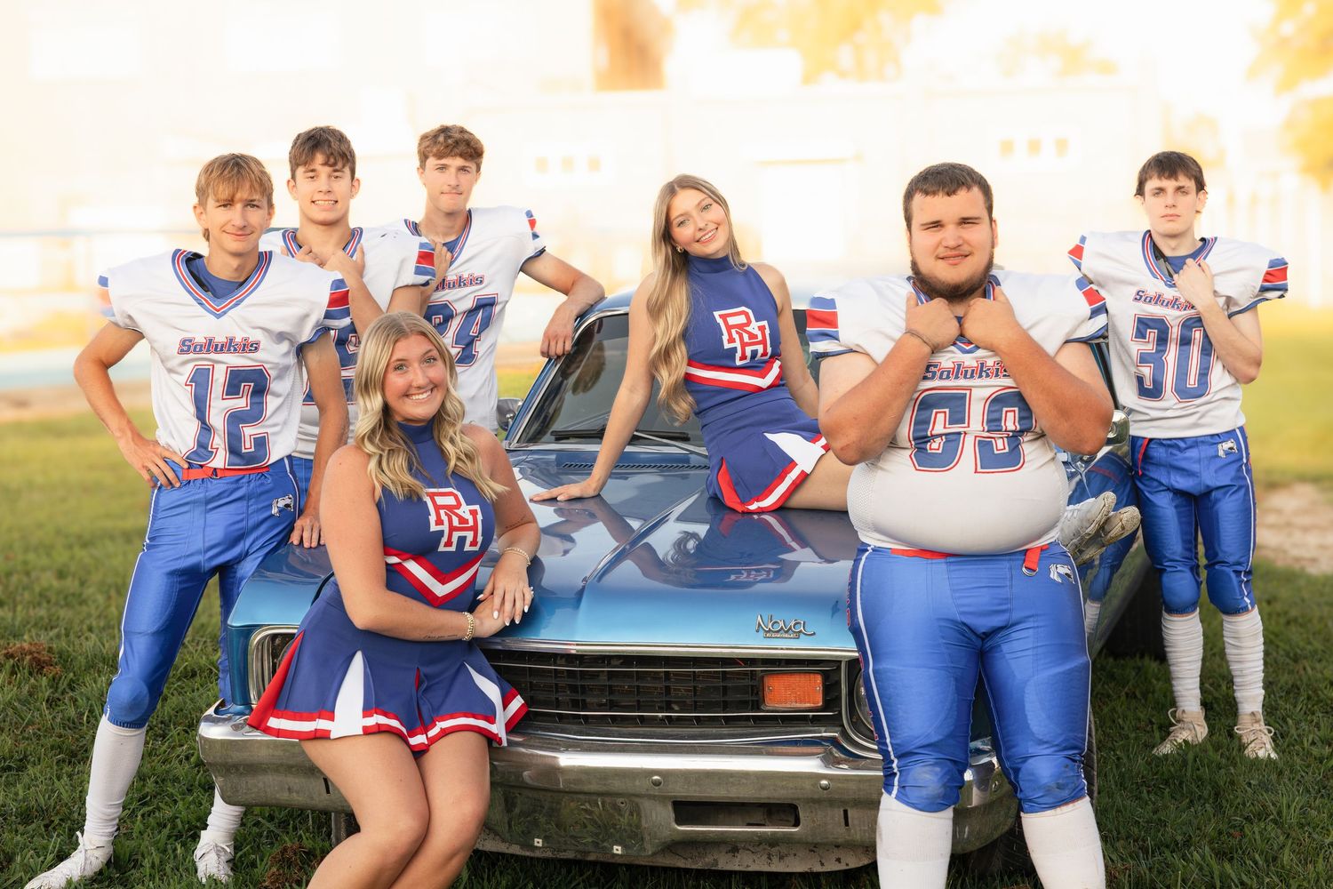 High school football players and cheerleaders pose together in front of a classic blue car on a football field.