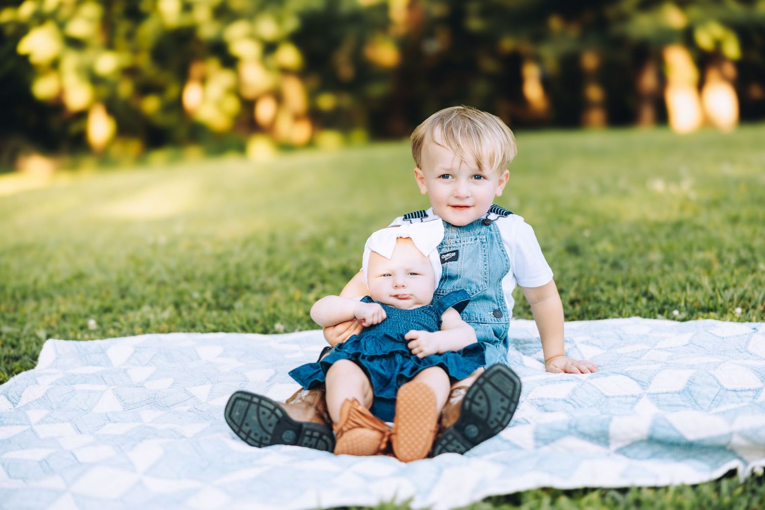 Two young siblings sit together on a blanket in a sunny green park, older toddler hugging baby sister.