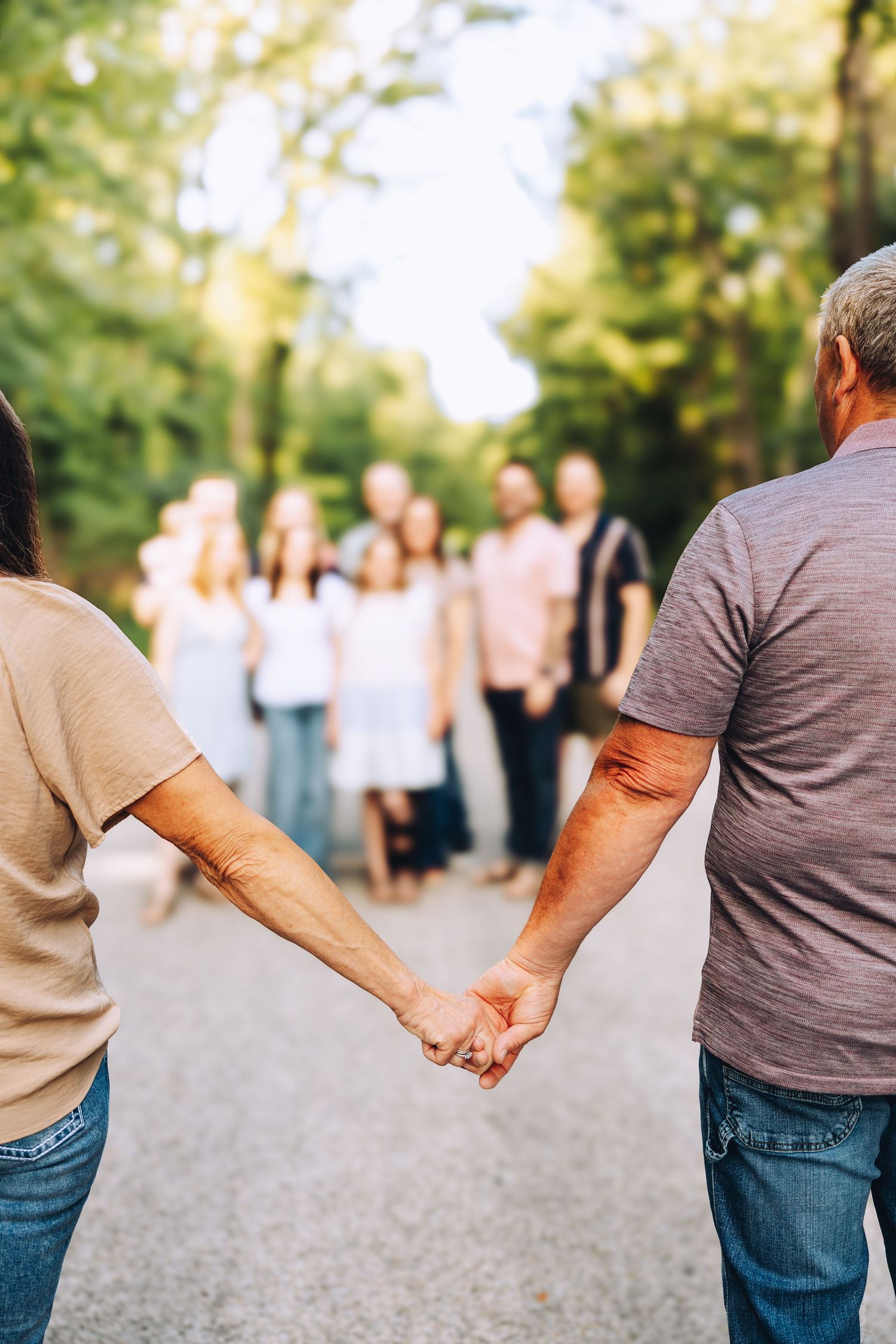 Couple holding hands on a tree-lined road with a large family group gathered ahead.