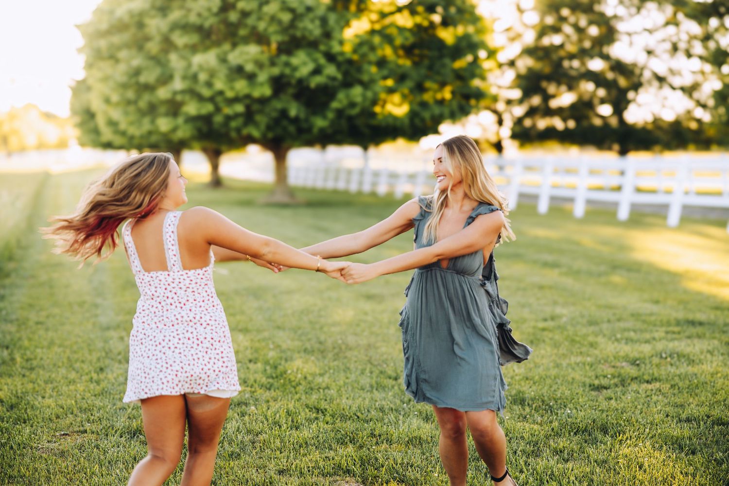 Two young women hold hands and spin joyfully on a green lawn at golden hour near a white fence.