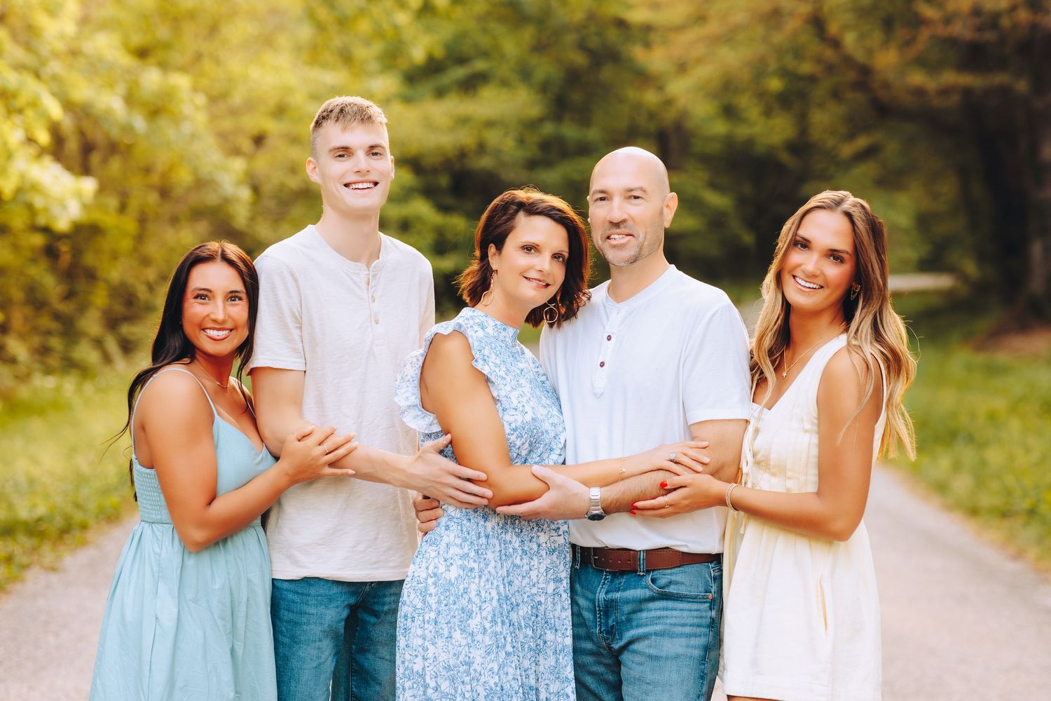 Happy family of five embracing on a scenic tree-lined path in warm golden summer light.