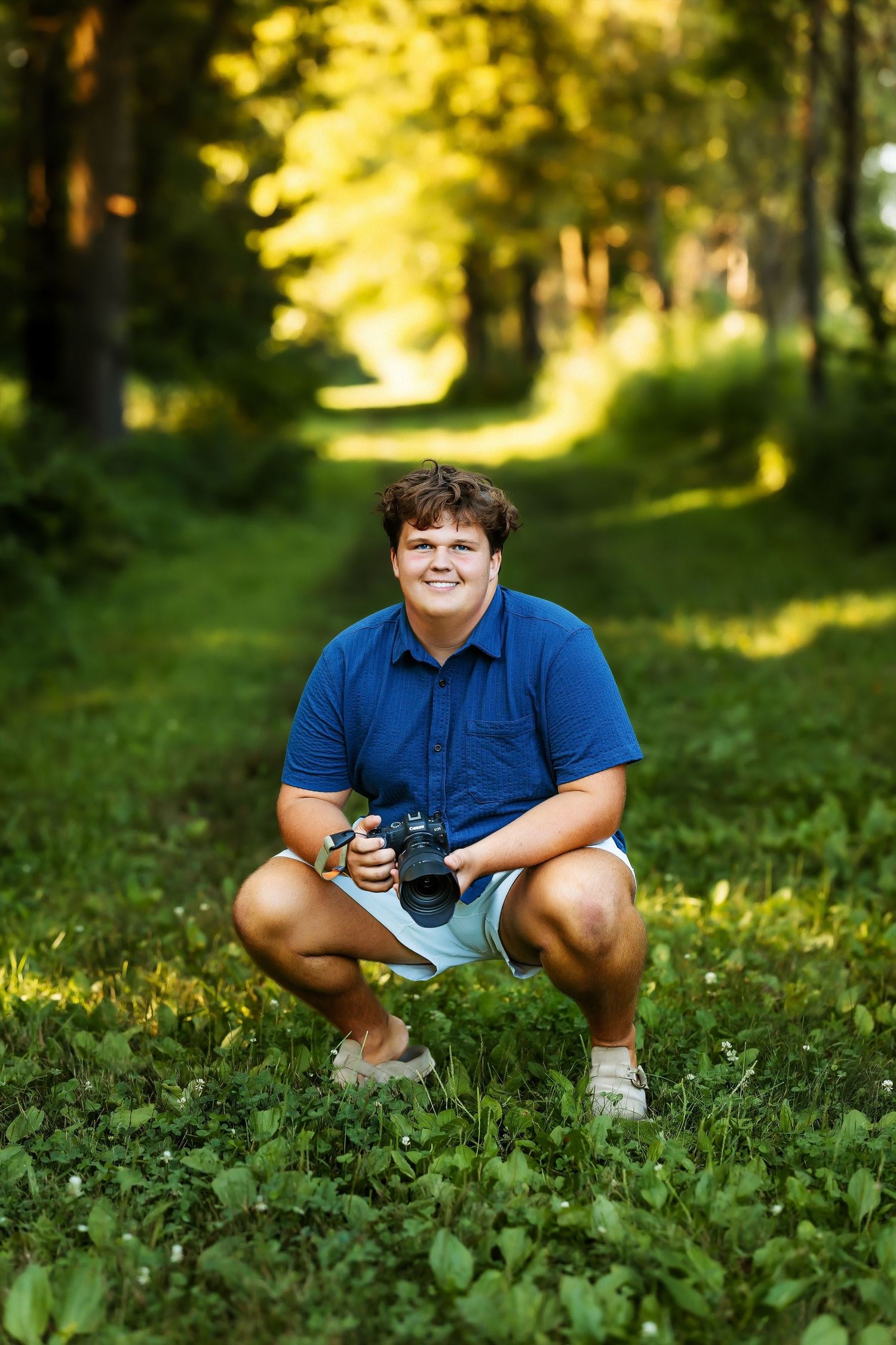 Smiling teen boy crouching in forest path holding a camera in bright green summer woods.