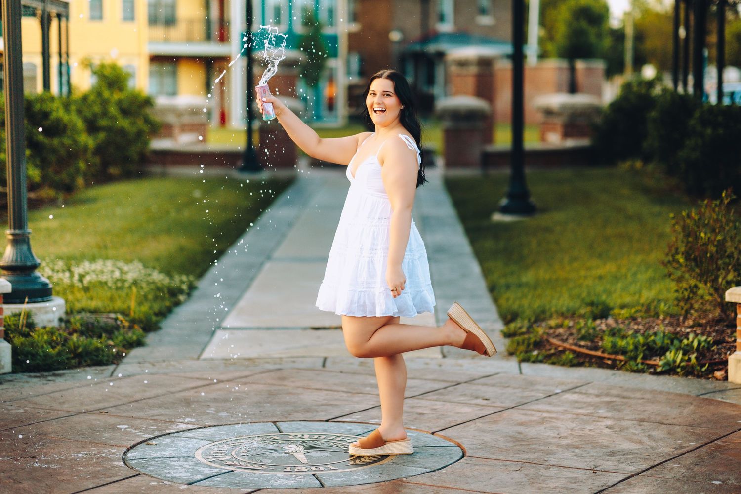 Young woman in white dress playfully splashing water fountain on a sunny outdoor plaza.