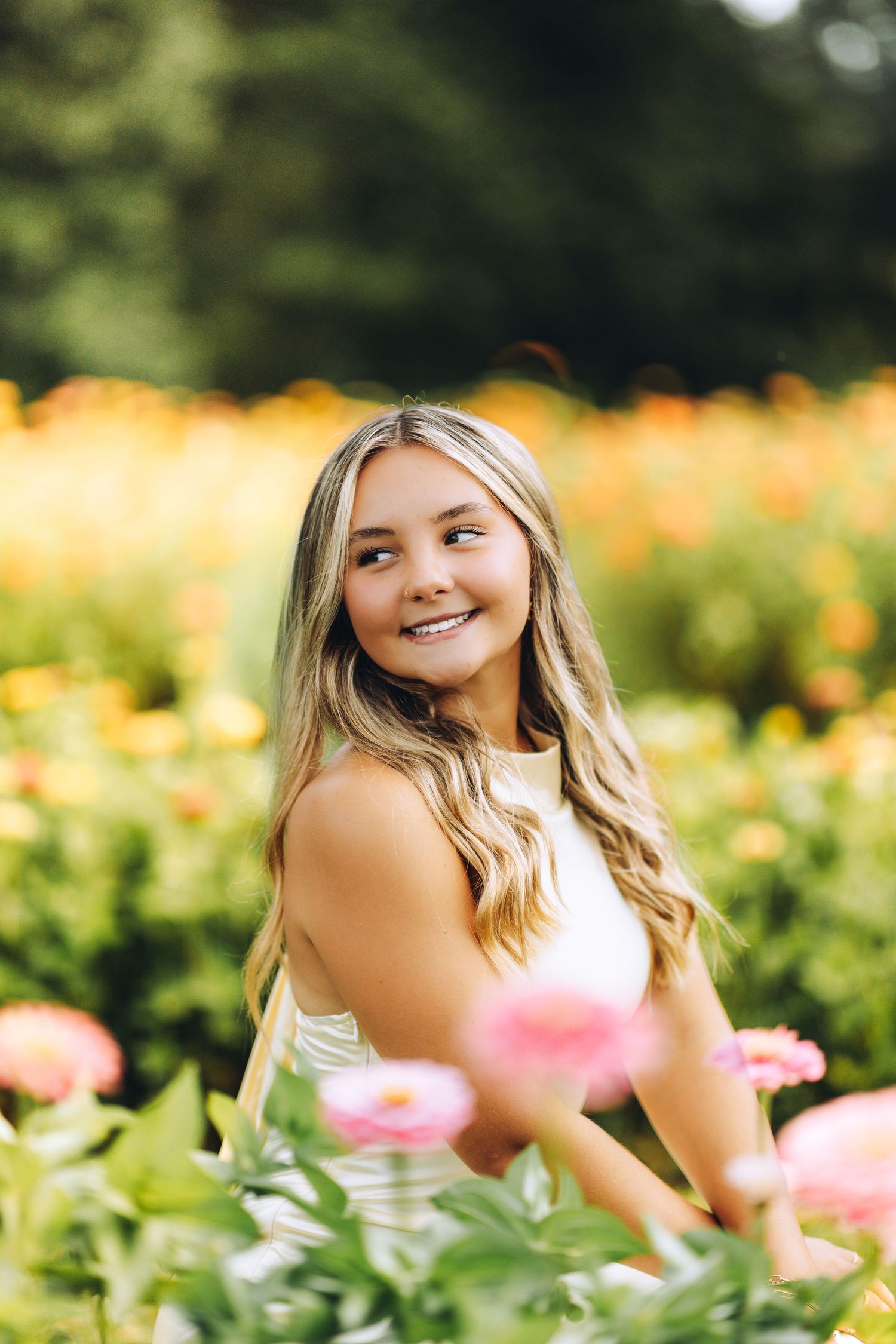 Smiling teenage girl with blonde hair posing in a colorful flower field with pink and yellow blooms.