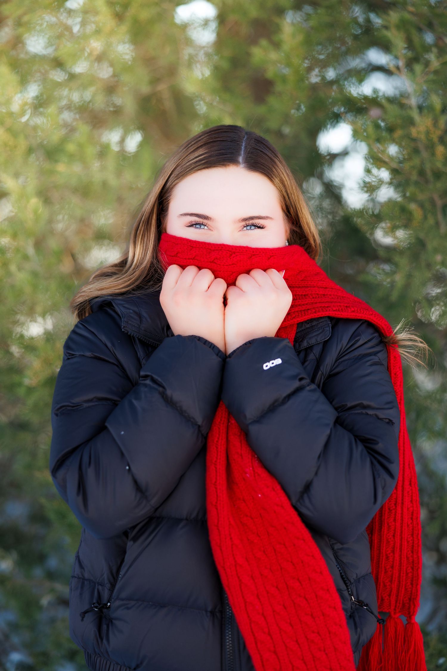 Teen girl with blue eyes peeks over a red knit scarf wearing a black puffer coat outdoors in winter.