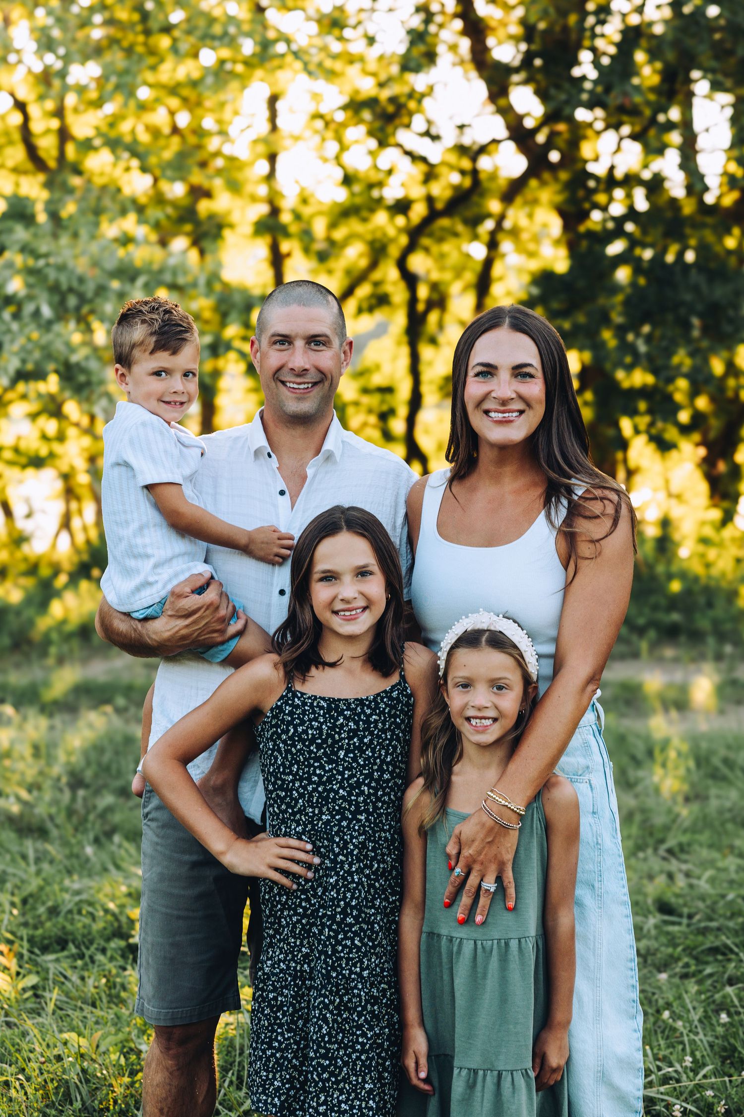 Happy family of five smiling together outdoors in a lush green sunlit park setting.