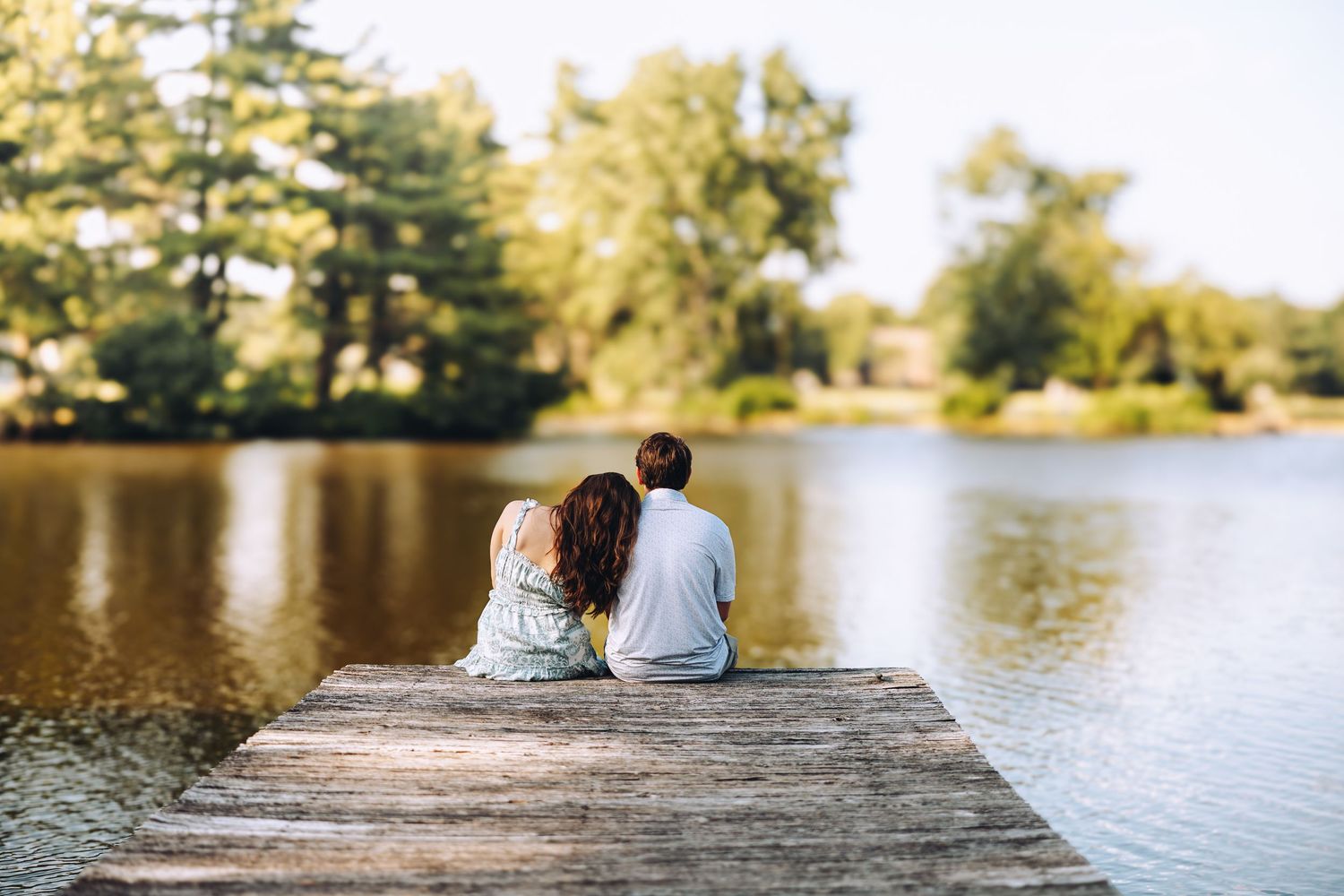 Couple sits together on a wooden dock overlooking a calm lake surrounded by lush green trees.