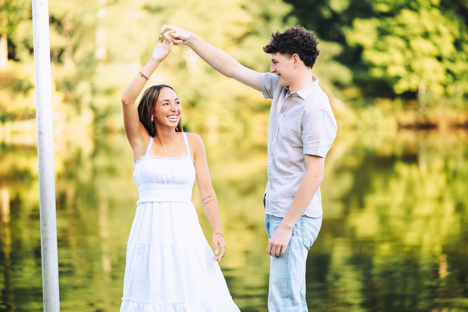 Happy couple dances outdoors near a lake, woman in white dress being twirled by man in casual attire.