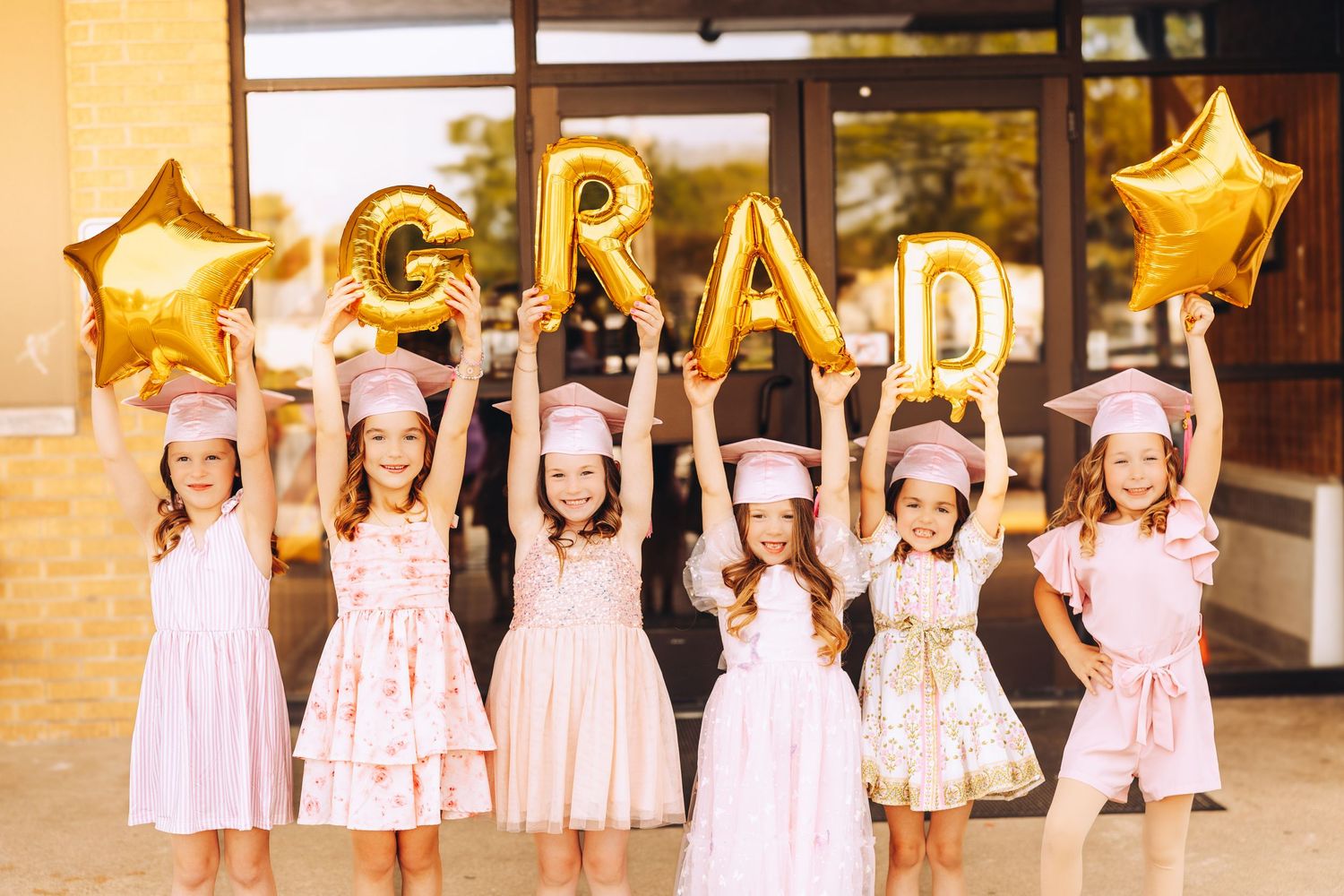 Six young girls in pink graduation caps holding gold GRAD balloon letters at a kindergarten ceremony.