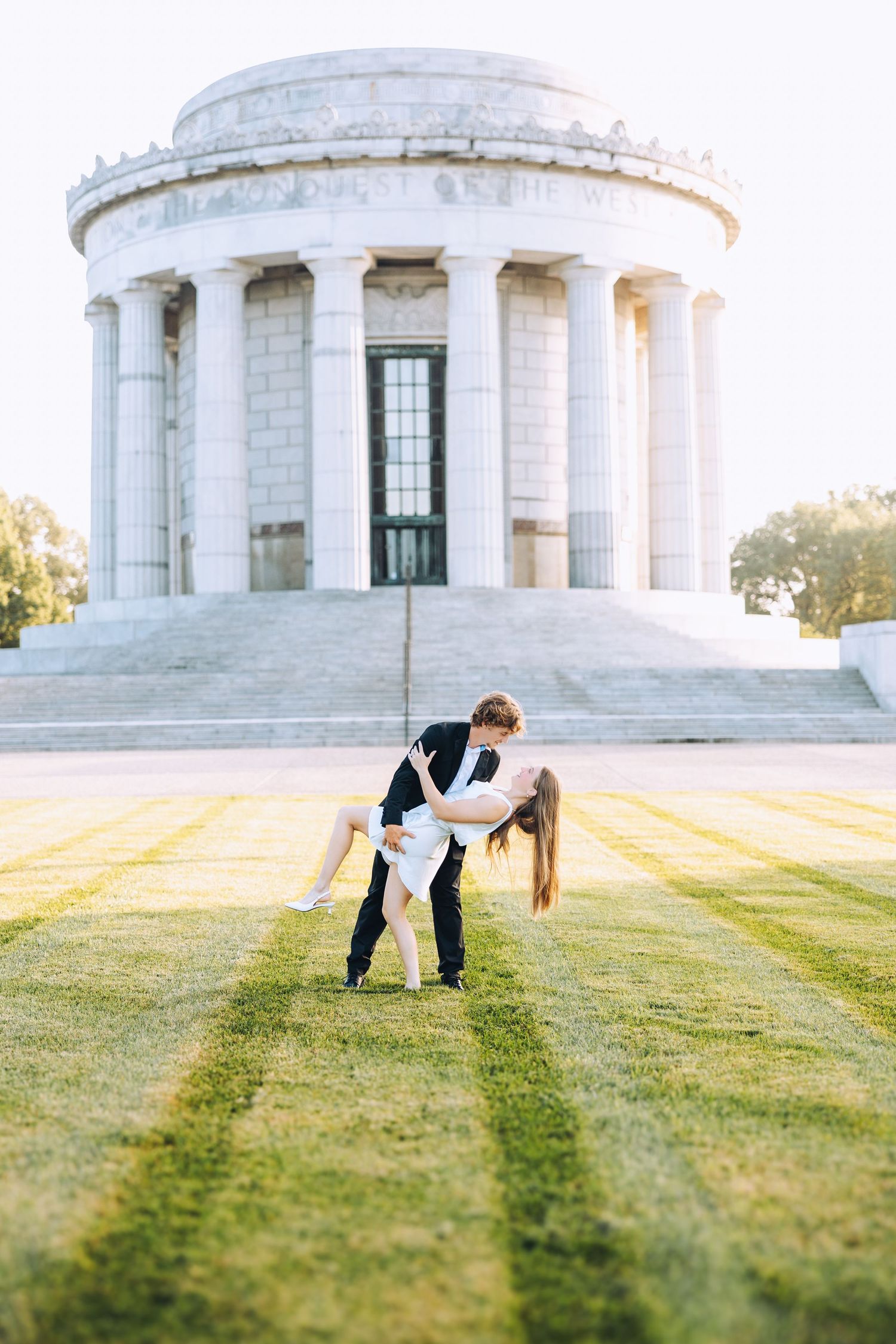 Couple sharing a romantic dip kiss on green lawn in front of a white neoclassical memorial monument.