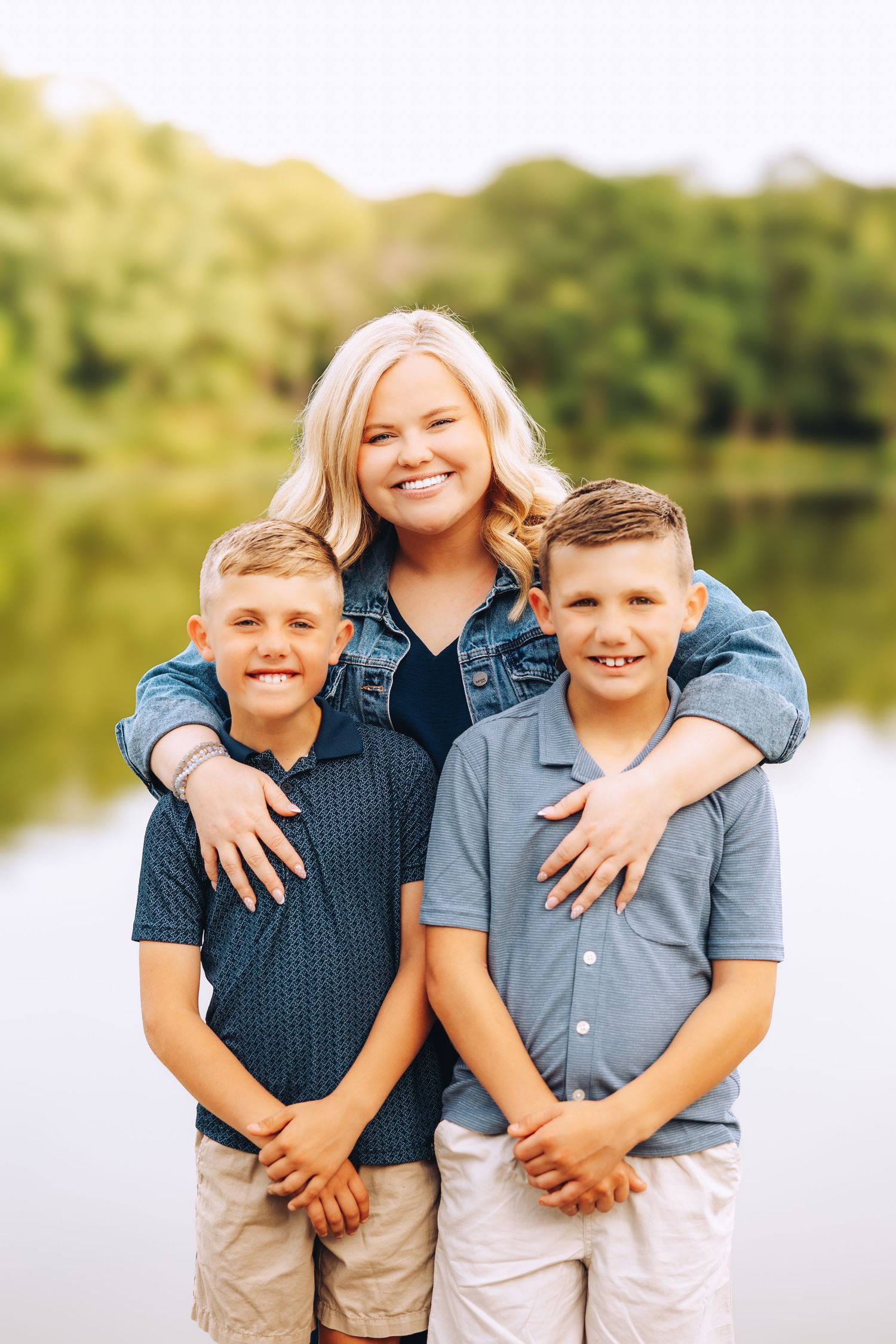 Smiling blonde mother hugging two young boys in blue shirts near a scenic lakeside.