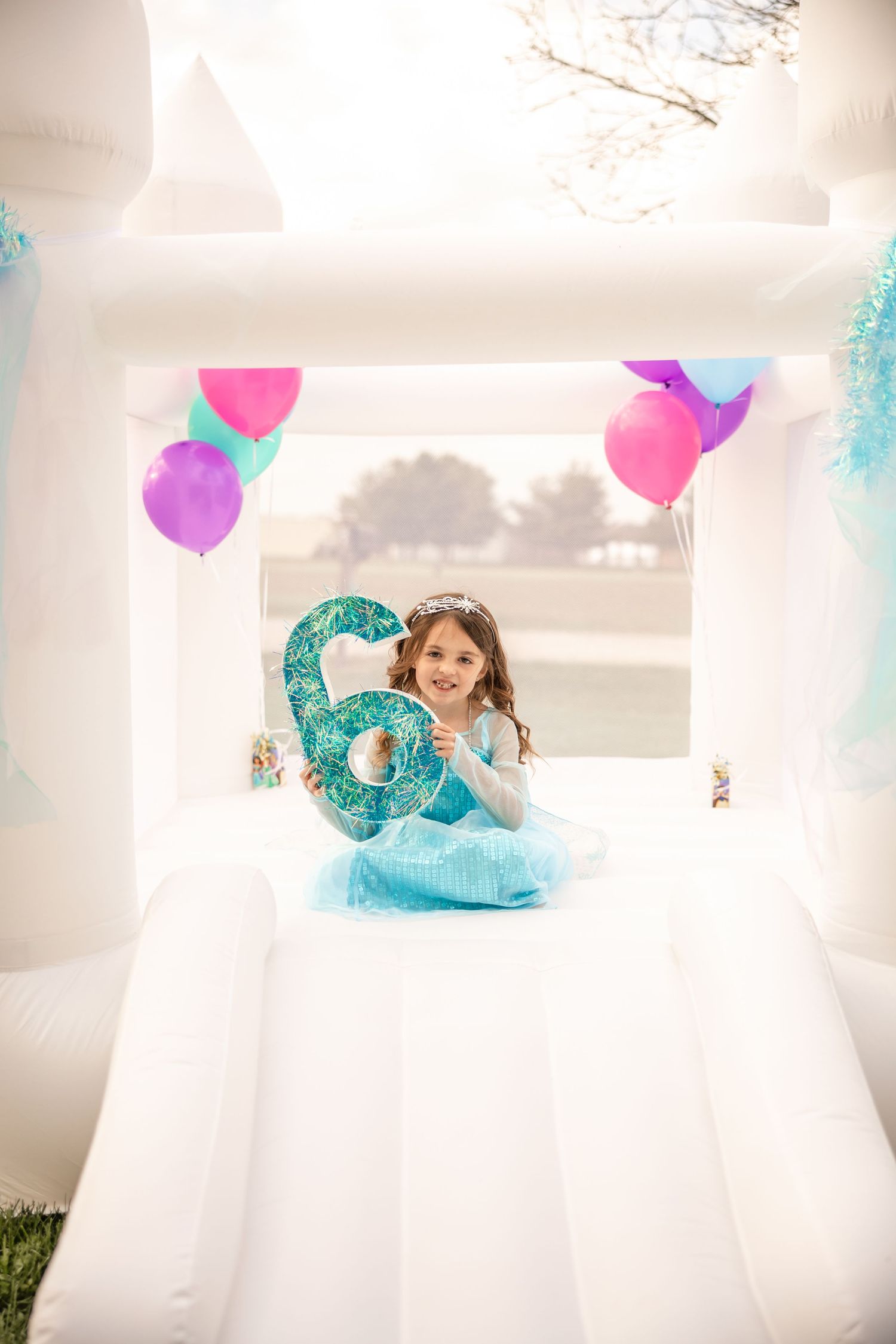 Young girl in Elsa costume holding number six balloon inside a white bounce house.