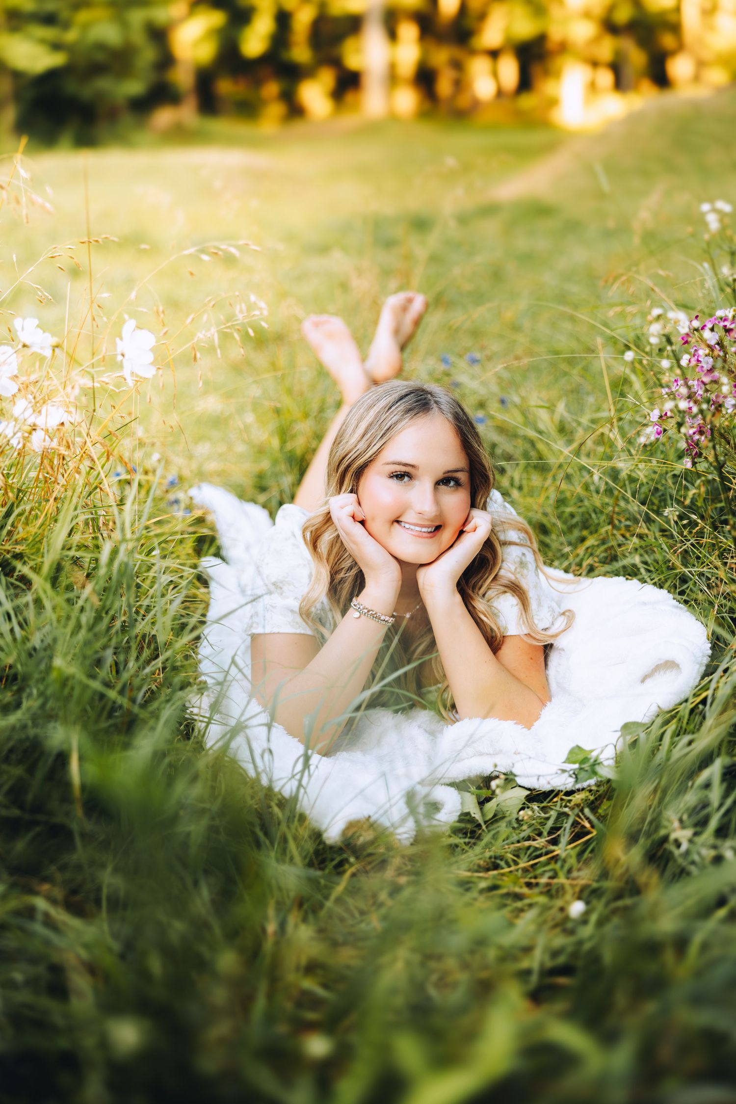 Smiling blonde teen girl in white dress lying in lush green meadow with wildflowers in sunlight.